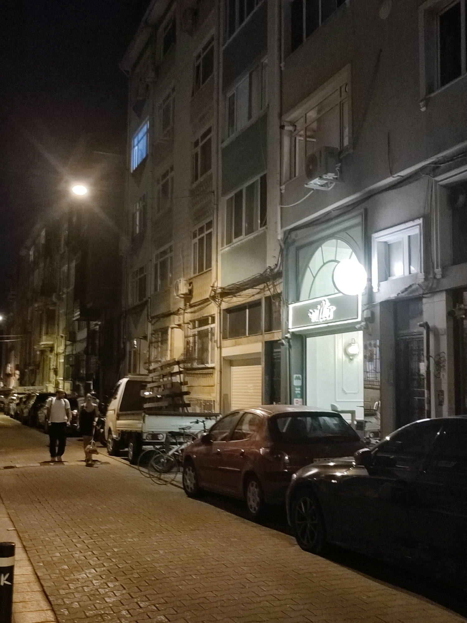 The image shows a street scene at night in Kadıköy, Turkey. A multi-story building with several windows lines the right side of the street. A lamppost with a bright light is visible on the left side of the street. Several cars are parked along the street, and a truck with a wooden rack is positioned in the middle. Two people are walking on the sidewalk, one walking a dog. A bicycle is leaning against the truck. The street is paved with cobblestones, and the overall lighting suggests it is nighttime. A business with illuminated signage is visible on the ground floor of the building.