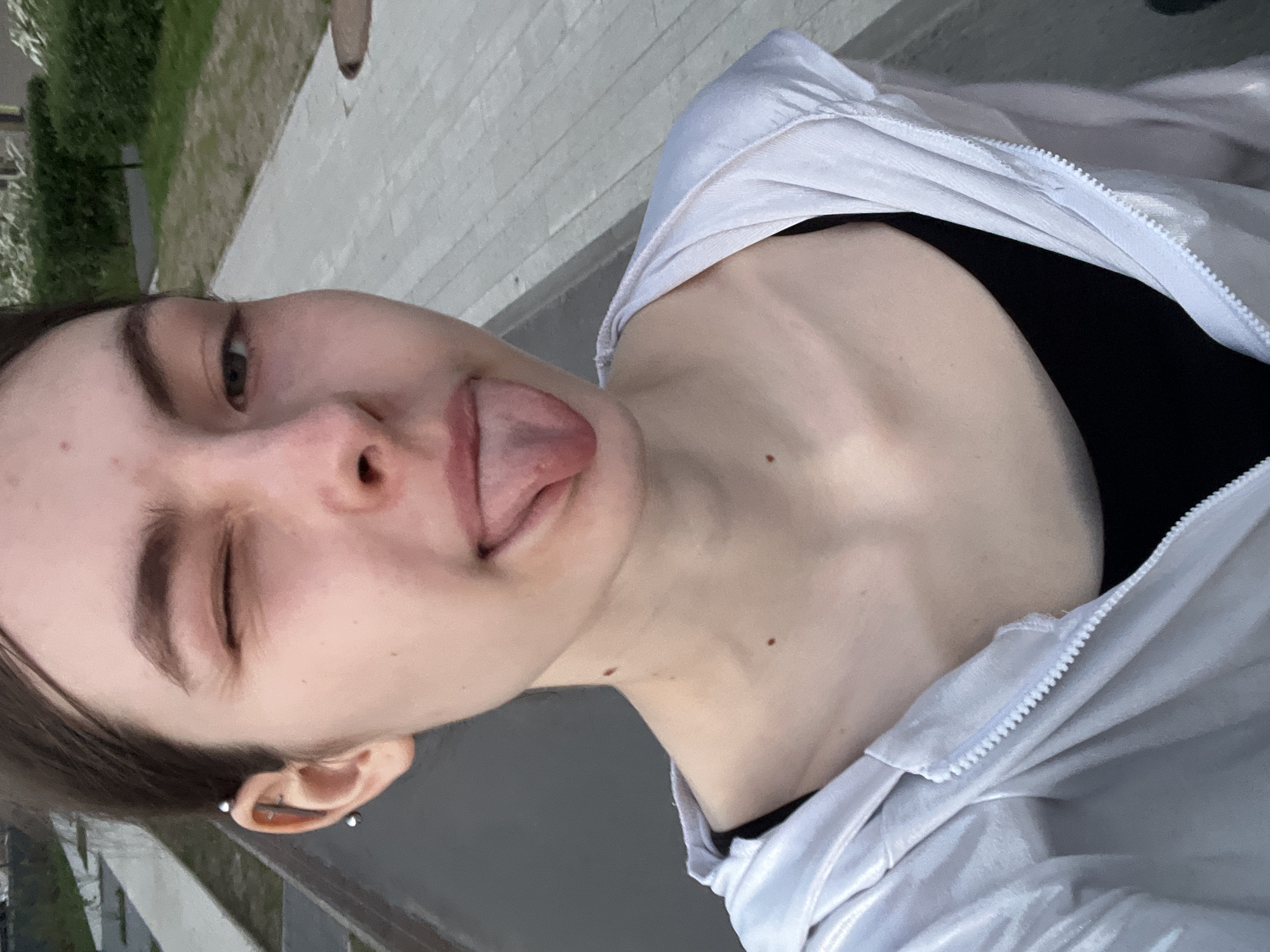 A young woman is taking a selfie outdoors. She is winking one eye and sticking out her tongue. She is wearing a white zip-up hoodie over a black top. She has a double helix earring in her left ear. The background shows a sidewalk and some greenery, suggesting an urban environment. It is not possible to confirm the specific location of Saint Petersburg from this image alone.