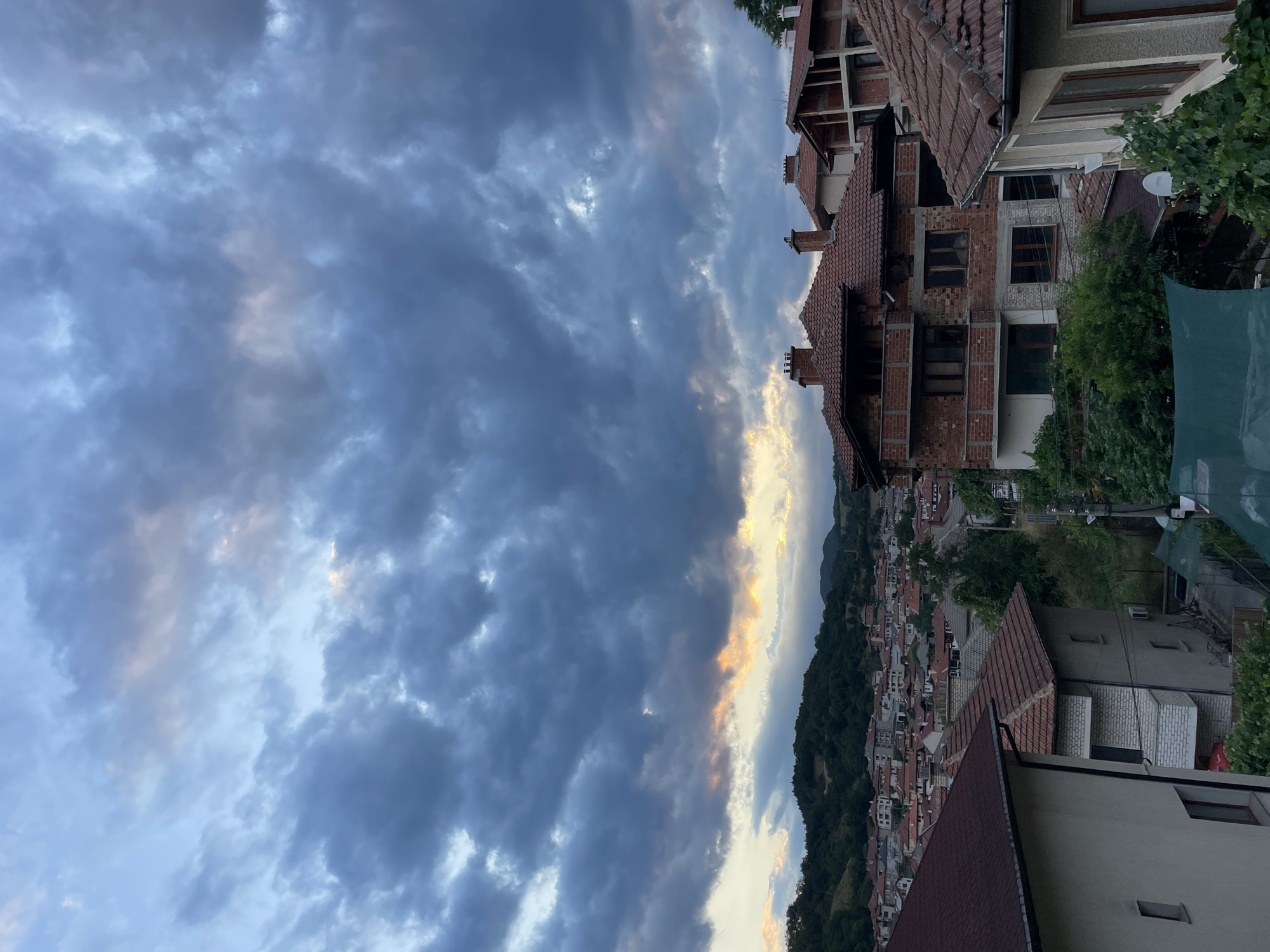 This is an outdoor, eye-level, vertical, full shot of a town with a cloudy sky in Zlatograd, Bulgaria. The clouds are dark and appear to be gathering, with some streaks of sunlight breaking through the gloom near the horizon. In the foreground, the roofs of several houses are visible. These houses are of varying architectural styles, with some appearing older and others under construction. The houses are predominantly made of brick and covered with terracotta roof tiles. Green foliage surrounds some of the buildings. In the background, a hillside covered in trees and more houses slopes upwards. The overall impression is of a town preparing for or experiencing a change in weather, with a dramatic sky overhead.