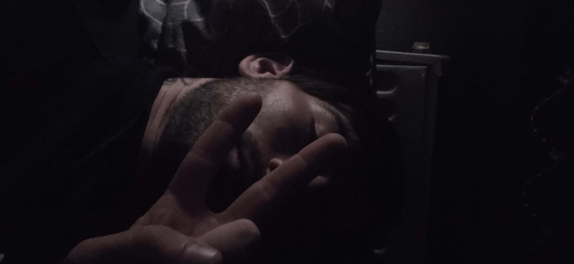 A person with dark hair and a beard is lying down, with their head resting on a patterned pillow. They are covering their face with their hand, showing two fingers extended in a peace sign. The lighting is very dim, with only a small portion of their face and hand illuminated. Behind the person, a dark, indistinct object with a reflective surface is visible. The background is predominantly black, suggesting a dark environment, possibly indoors at night.