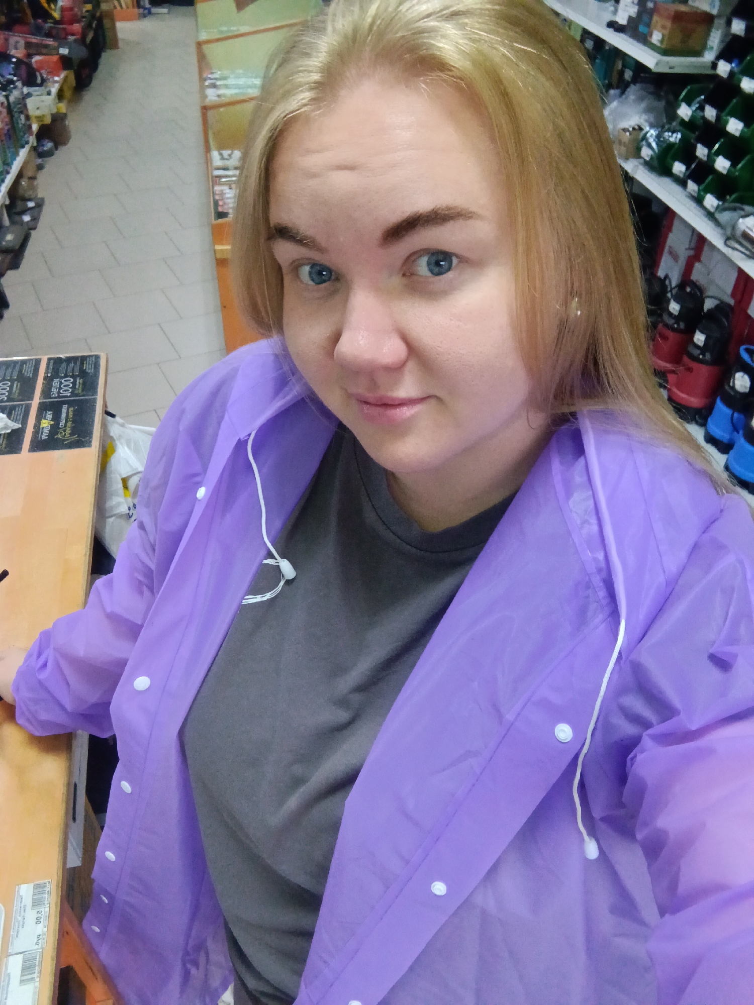 A young Caucasian woman with blonde hair and blue eyes is taking a selfie in a retail store. She is wearing a grey t-shirt and a translucent purple raincoat. The background shows shelves stocked with various products, including what appear to be bottles and containers. The floor is tiled, and a wooden counter is visible to her left. The scene is brightly lit, suggesting an indoor environment. The woman is looking directly at the camera with a slight smile. The retail store appears to be located in the city of Fokino, Russia, as suggested by the context of the request.