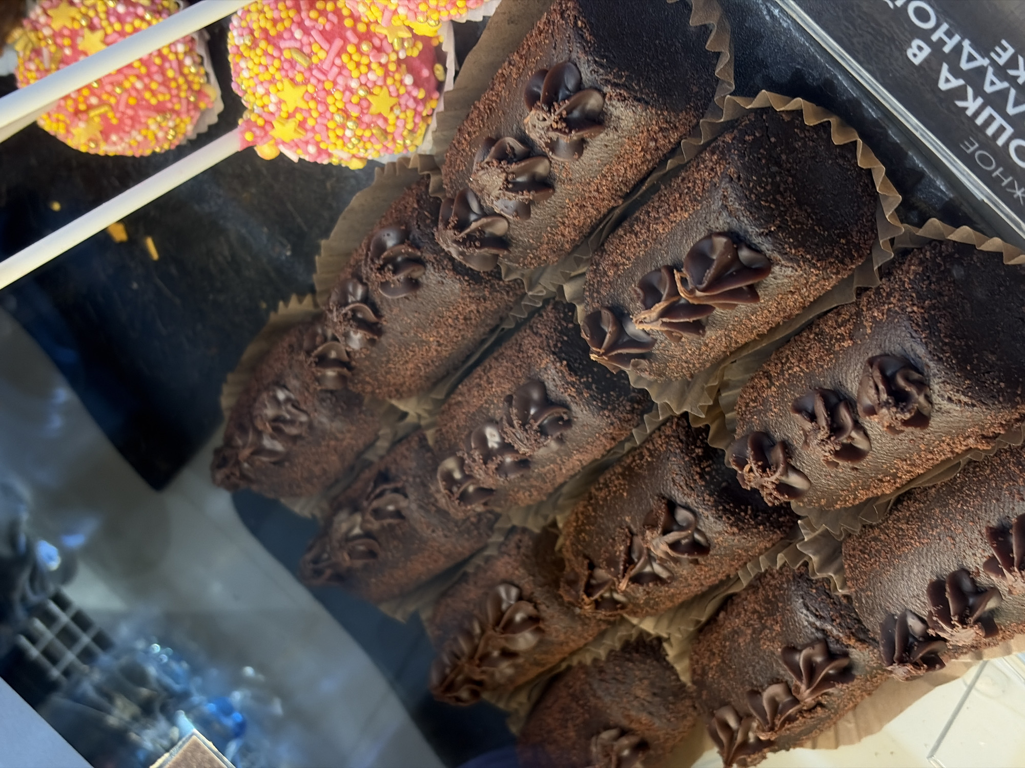 The image shows a close-up, high-angle view of a display case filled with assorted pastries. In the foreground, numerous chocolate-coated cylindrical cakes, dusted with cocoa powder and topped with chocolate frosting in a floral pattern, are arranged in paper liners. To the right and slightly further back, two cake pops decorated with pink frosting and a colorful assortment of sprinkles and yellow star-shaped candies are visible, each on a white stick. The background is blurred but suggests a retail environment, possibly a bakery or confectionery shop. Text visible at the bottom right, partially obscured, appears to be in Cyrillic. Based on the visible text, this scene is located in Nikolskoye, Russia.