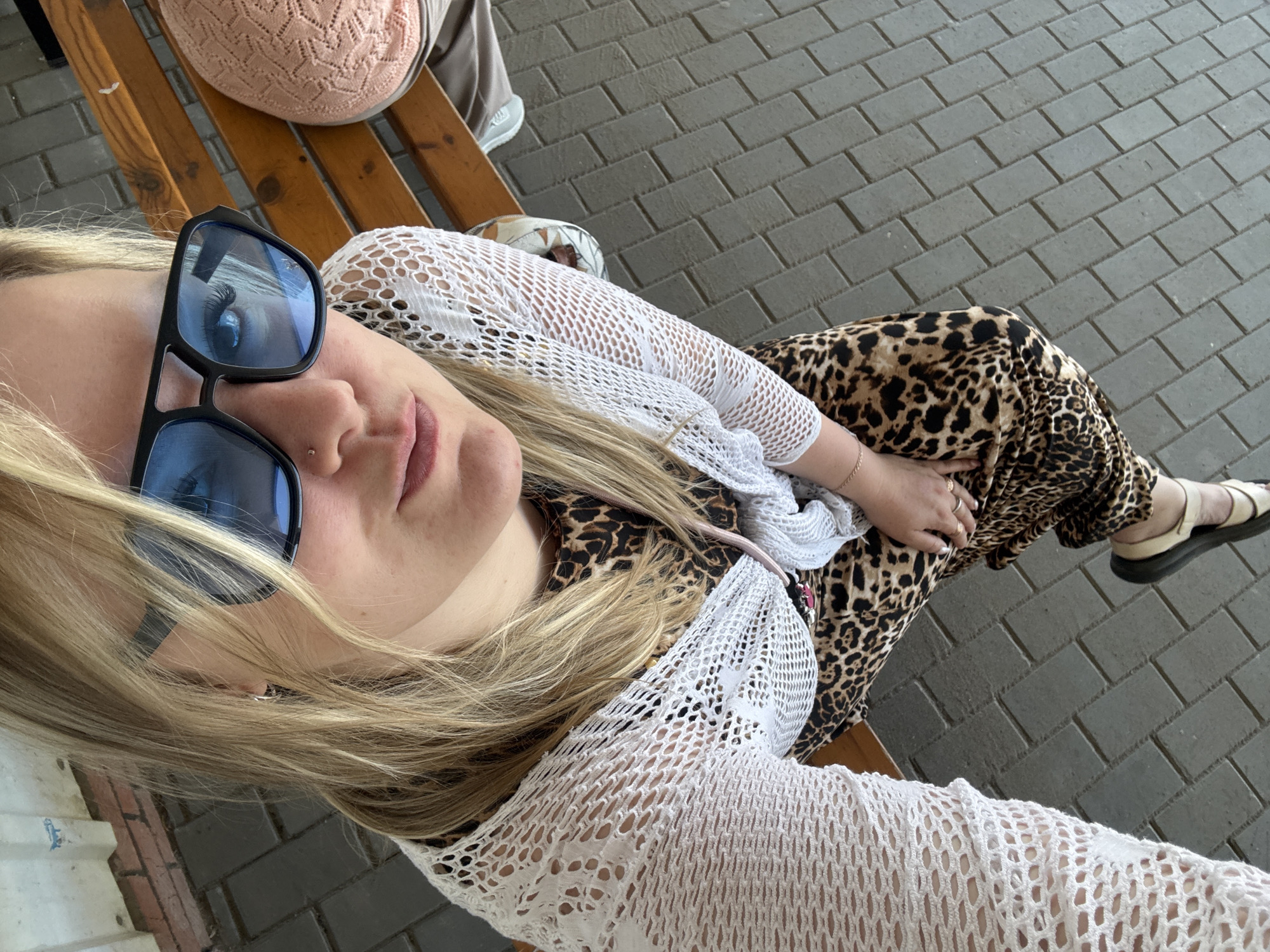 A medium shot depicts a fair-skinned woman with blonde hair, wearing sunglasses, a white crocheted top, and a leopard-print dress, reclining on a wooden bench. She has a nose piercing. The ground is tiled with gray bricks. Another person wearing a pink knit top and light-colored pants is partially visible in the top left corner. The location is Chaikovsky, Russia.