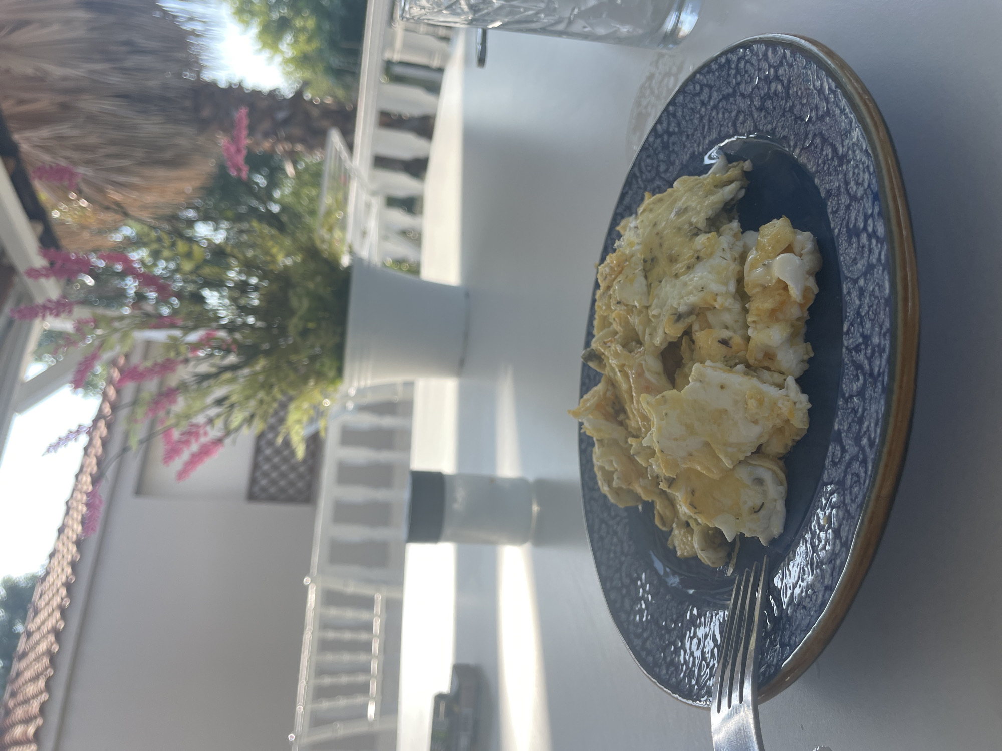 A close-up shot captures a plate of scrambled eggs on a textured blue plate with a dark rim, set on a white table. A fork rests on the left side of the plate. In the background, a salt shaker with a black lid is visible on the table. Beyond that, a blurred potted plant with pink flowers and a palm tree are present, suggesting an outdoor setting. White railing and a tiled roof are also visible, hinting at a building or structure. The environment appears to be a sunny outdoor dining area, possibly at a cafe or restaurant in Skala Avdiron, Greece, characterized by its Mediterranean climate and coastal proximity.