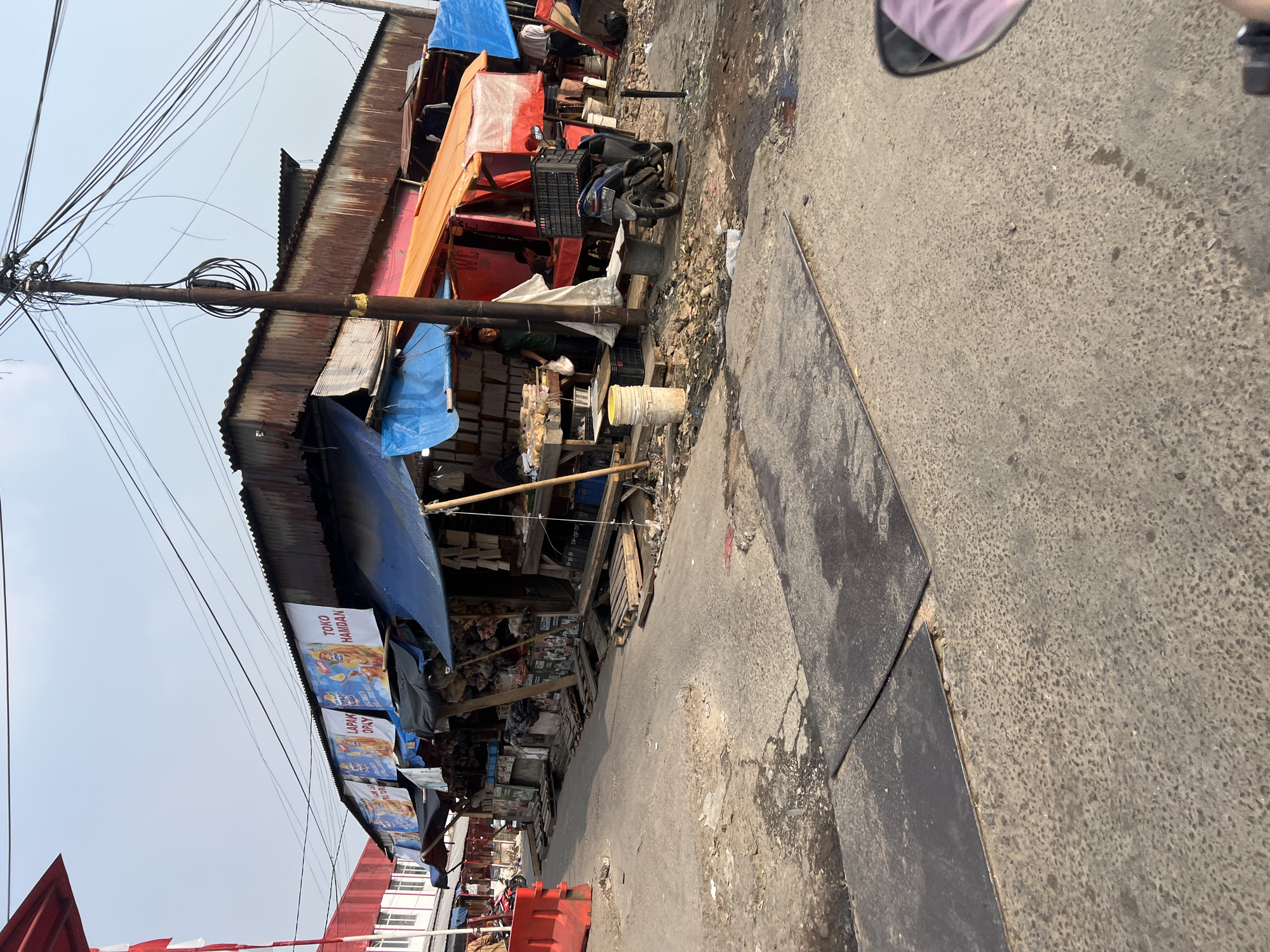 This is a street-level, medium-distance, eye-level, outdoor, daytime, photographic image of a market scene in Citeureup, Indonesia. The image captures a row of market stalls with corrugated metal roofs, some covered with tarpaulins in blue and orange. The stalls are lined up along a dirty, paved street, with debris and puddles visible on the ground. A person in a green shirt is standing near a stall displaying food items on a table. A motorcycle is parked in front of one of the stalls, and a red traffic barrier is visible on the left side of the image. Several banners are hanging from the stalls, one of which reads "TOKO HAMDAN". Overhead, a tangled network of electrical wires and a utility pole are prominent. The sky is clear and bright, indicating a sunny day.