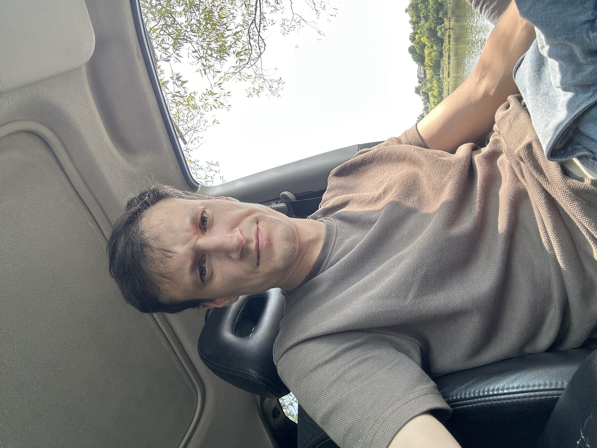 A man with short, dark hair and light skin is seated in the driver's seat of a car. He is wearing a grey, short-sleeved, textured t-shirt and blue jeans. The car's interior is visible, including a black leather seat and a sunroof. Outside the car, a body of water is visible, with trees and houses on the opposite bank. The sky is bright and overcast. The location is the city of Makiv, Ukraine.