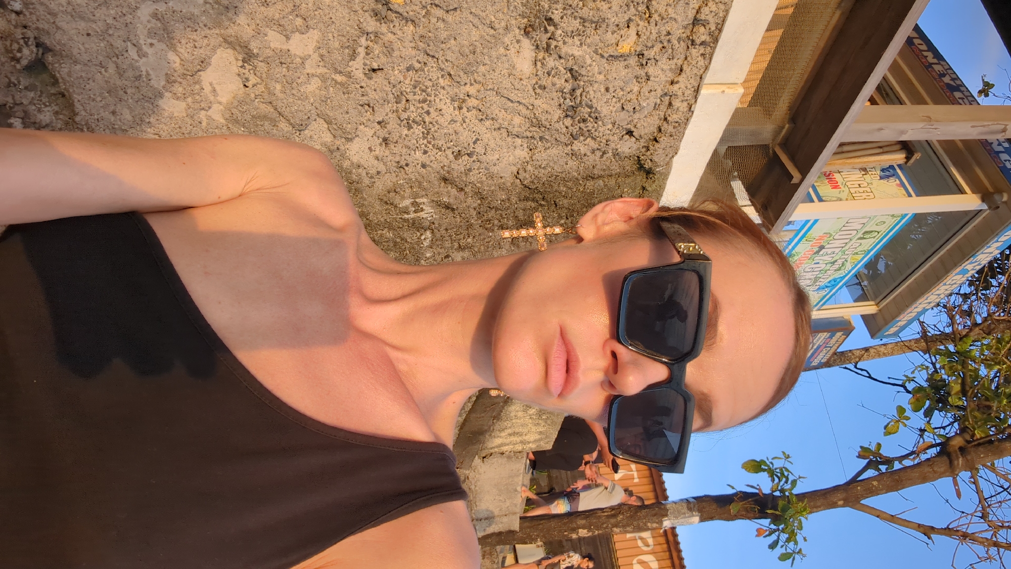 The media file shows a woman in a black tank top, wearing large black sunglasses and a bejeweled cross earring. She is lying down on a rough, textured surface, possibly a stone wall, with sunlight casting a warm glow on her skin. The setting appears to be outdoors, with a clear blue sky visible in the background. To her right, there is a wooden structure with a sign that reads "MONEY CHANGER" in large letters. The sign also features images of currency. A tree branch with green leaves extends into the frame from the right. The lighting suggests it is either sunrise or sunset. The overall mood is relaxed and casual, possibly indicating a vacation or leisure activity in a tropical location.