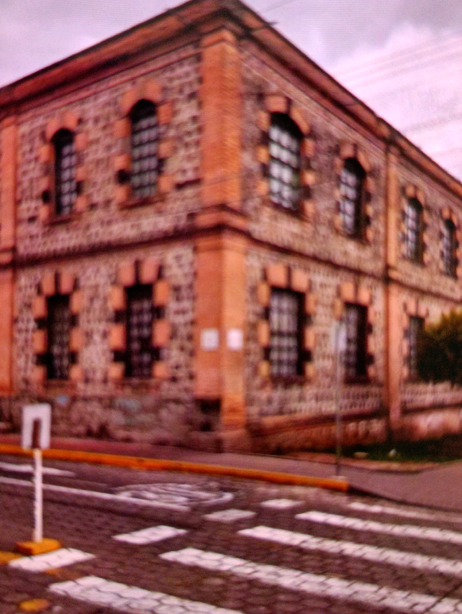The image shows a blurred view of an old, brick building with red-orange trim. The building has multiple arched windows with dark panes and decorative brickwork. In the foreground, a crosswalk is visible on a cobblestone street, with a white signpost on a yellow base to the left. The sky is a pale pinkish-purple, suggesting it might be dawn or dusk, or an effect of the camera or lighting. The overall scene depicts a quiet urban environment, possibly in a historic district, though no specific landmarks are identifiable in the provided view. The location is not explicitly stated in the image itself, but it has been identified as San Roque, Ecuador.