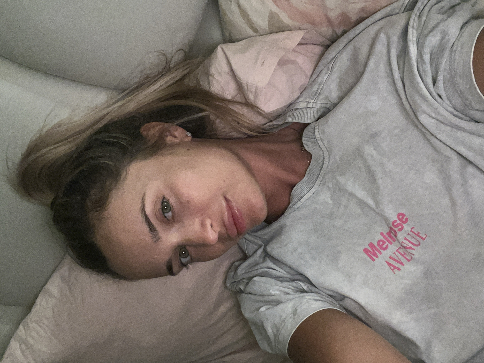 A woman with blonde hair pulled into a high ponytail, wearing a light gray t-shirt with pink text that reads "Melrose Avenue," is lying on a bed. Her eyes are green, and she has light skin. She is looking directly at the camera. The background consists of pillows, one of which is pink. The lighting suggests an indoor setting, possibly at night or in a dimly lit room. There is no information in the image to suggest this scene is located in Sharm El Sheikh, Egypt.