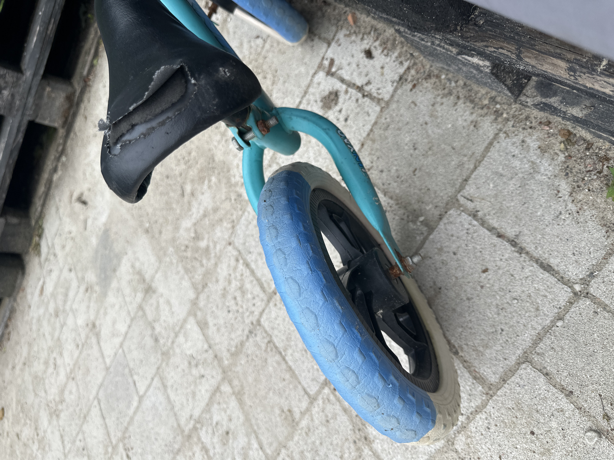 The image shows a close-up view of a blue balance bike with a worn black seat, resting on a light-colored paved area. The bike's front tire is blue with a white rim. The background features a dark wooden structure, possibly a fence or bench, and some greenery. The scene appears to be outdoors, possibly in a residential area or park, in the city of Malinovka, Russia.