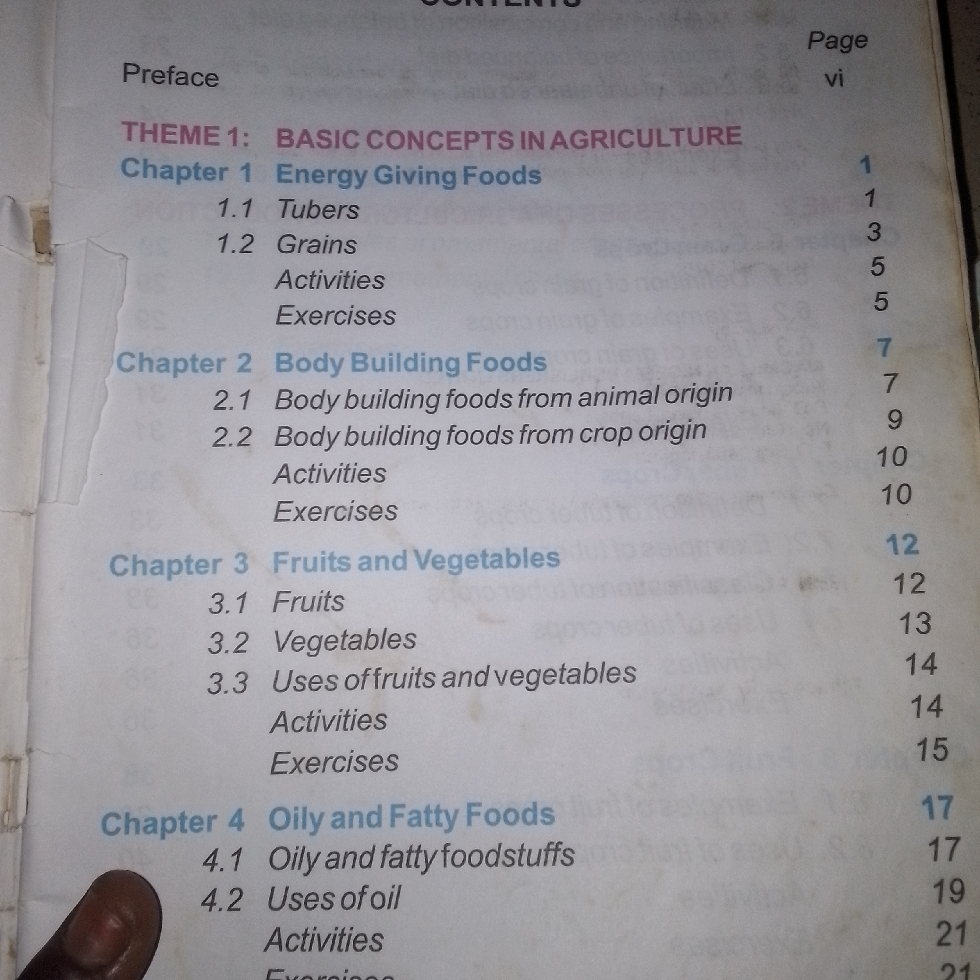 The image shows a page from a book's table of contents. The text is in English and is organized by chapters and themes, with corresponding page numbers listed on the right side of the page. The left side of the page is largely blank due to a tear or damage to the paper.
The visible content includes:
- "Preface" followed by a page number "vi".
- "THEME 1: BASIC CONCEPTS IN AGRICULTURE"
- "Chapter 1 Energy Giving Foods" with subtopics "1.1 Tubers", "1.2 Grains", "Activities", and "Exercises", followed by page numbers 1, 1, 3, 5, and 5 respectively.
- "Chapter 2 Body Building Foods" with subtopics "2.1 Body building foods from animal origin", "2.2 Body building foods from crop origin", "Activities", and "Exercises", followed by page numbers 7, 7, 9, 10, and 10 respectively.
- "Chapter 3 Fruits and Vegetables" with subtopics "3.1 Fruits", "3.2 Vegetables", "3.3 Uses of fruits and vegetables", "Activities", and "Exercises", followed by page numbers 12, 12, 13, 14, and 14 respectively.
- "Chapter 4 Oily and Fatty Foods" with subtopics "4.1 Oily and fatty foodstuffs", "4.2 Uses of oil", "Activities", and "Exercises", followed by page numbers 15, 17, 17, 19, and 21 respectively.
The lighting suggests the image was taken indoors, possibly with flash photography. There is a thumb visible at the bottom left of the image, indicating someone is holding the page. The overall background is a light-colored paper. No location in a city is discernible from the image.