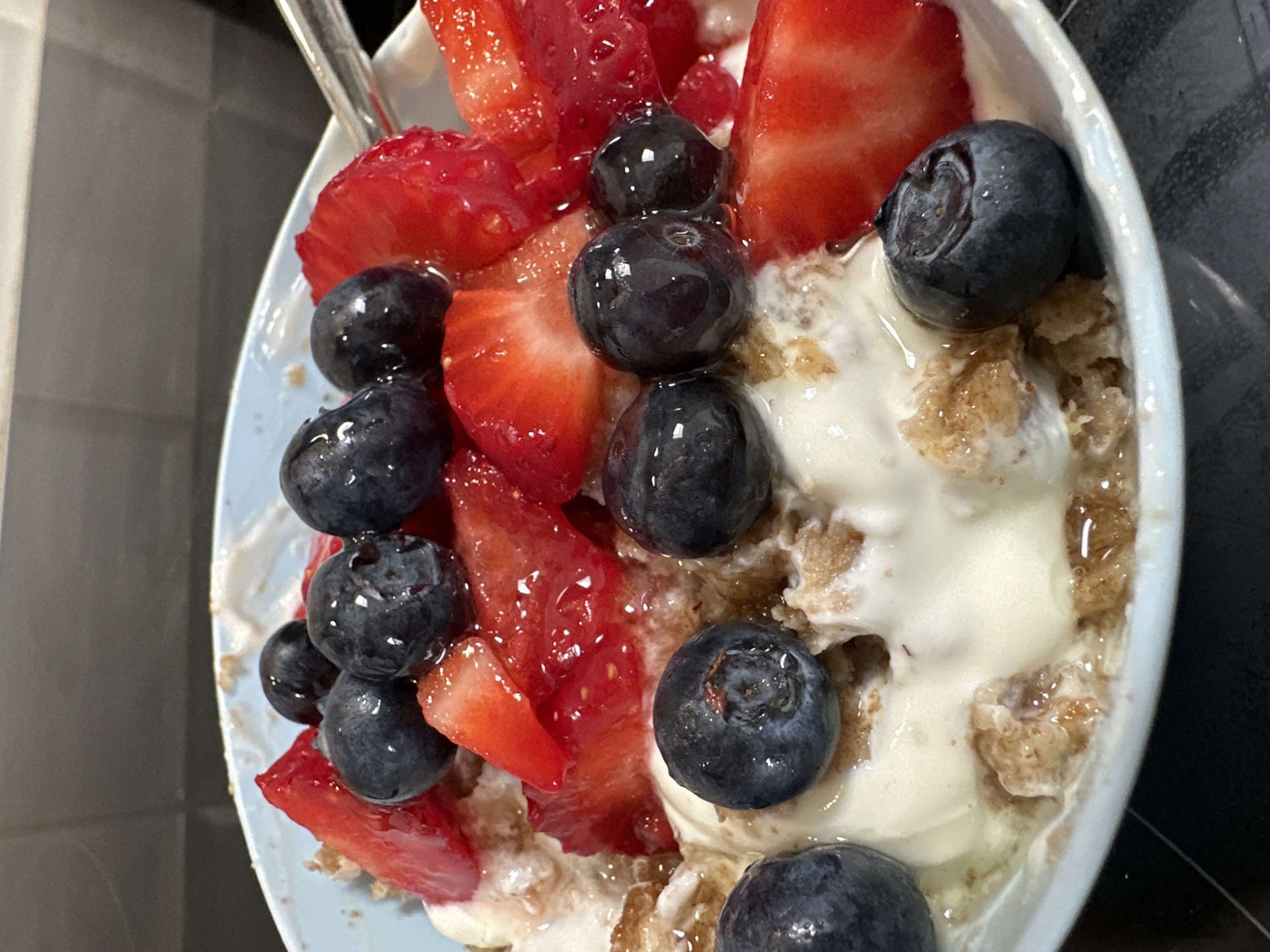 In Moortown, Ireland, a close-up shot captures a bowl of granola topped with fresh strawberries and blueberries. The bowl, a pale blue, rests on a dark surface. The granola appears to be mixed with a creamy white substance, likely yogurt or ice cream. Sliced strawberries, vibrant red, are interspersed with plump blueberries, their deep blue skins glistening. The lighting highlights the textures of the fruit and granola, creating a visually appealing still life. The background is blurred, showing a dark, textured surface that could be a countertop or table. A sliver of a metallic spoon is visible on the right side of the frame.