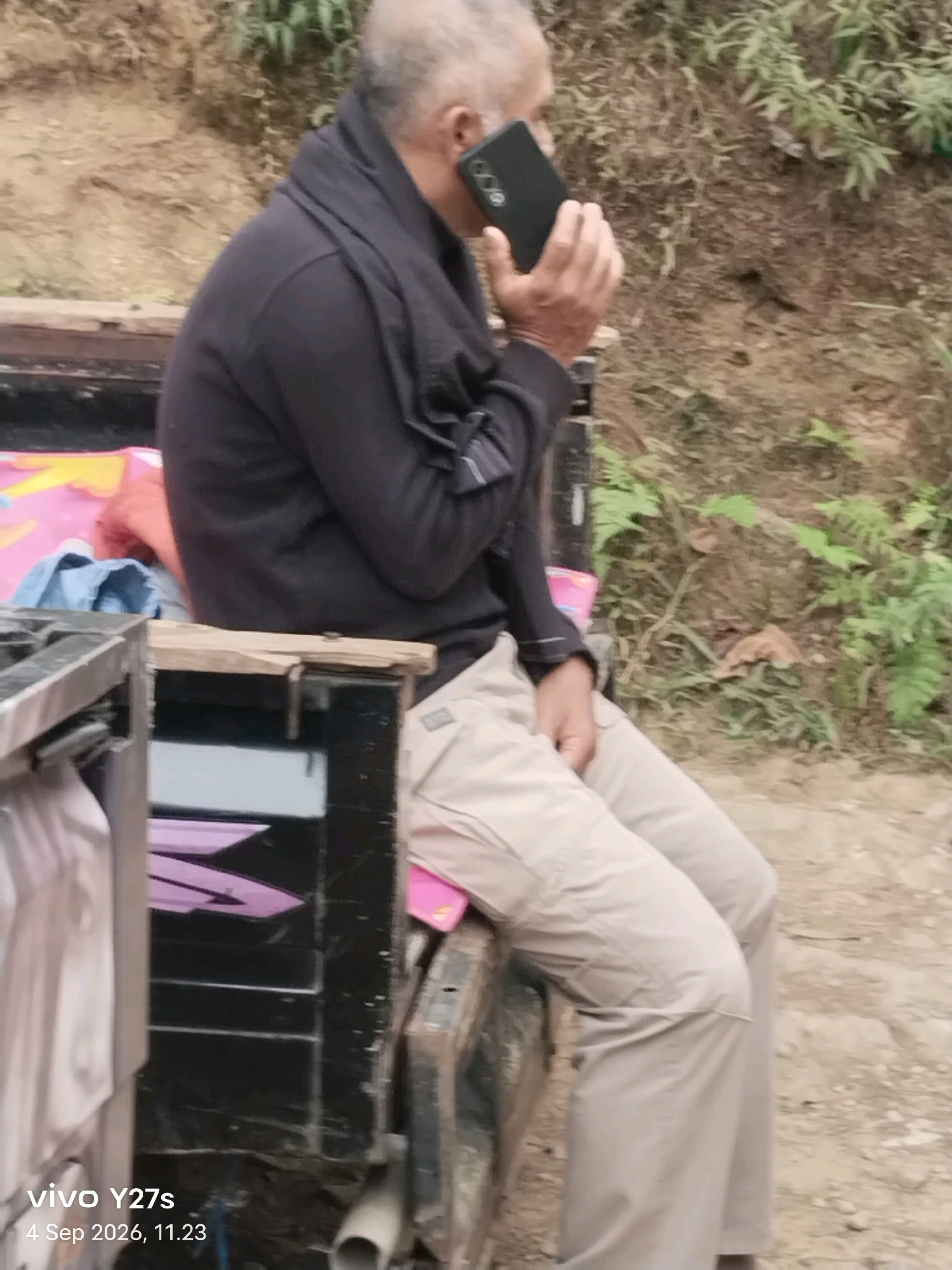 The image depicts a man sitting on the back of a vehicle, possibly a truck, and speaking on a mobile phone. He is wearing a dark long-sleeved shirt and light-colored pants. The vehicle appears to be parked outdoors, with a natural, somewhat rugged environment in the background, characterized by dirt, vegetation, and a hillside. The man's posture and the way he holds the phone suggest he is engaged in a conversation. The timestamp on the image indicates it was taken on September 4, 2026, at 11:23 AM. While the location is stated as Maron, Indonesia, there are no specific identifiable landmarks within the image that would pinpoint a precise location within the city or its immediate surroundings. The scene is captured from a side profile, focusing on the man and the immediate area around him.