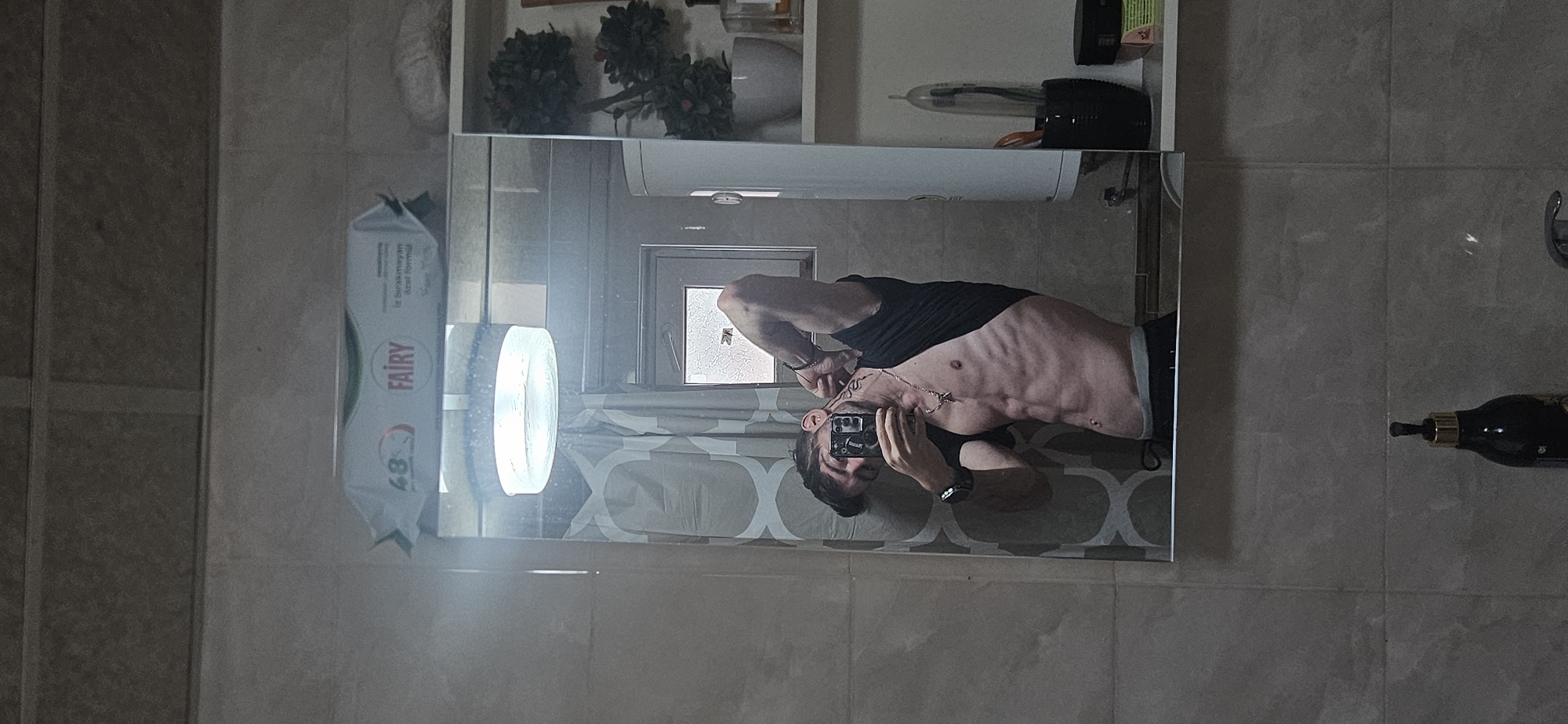 A man is depicted taking a mirror selfie in what appears to be a bathroom. The image is oriented vertically, with the man's head towards the bottom left and his feet towards the top right.
**Subjects:** The primary subject is a man with short, dark hair and a beard or stubble. He is wearing a black t-shirt pulled up to reveal his lean, muscular torso, showcasing visible abdominal muscles and ribs. He wears a smartwatch on his left wrist and a silver-colored necklace with a pendant around his neck. He is holding a smartphone to capture his reflection.
**Objects:** The central feature is a large, rectangular mirror reflecting the man and parts of the room. Above the mirror, a white shelf holds decorative green plants, a white ceramic container, and a black holder with what appears to be toothbrushes or similar bathroom accessories. To the left of the man's reflection, a white packet labeled "FAIRY" is visible, along with a bright, round object, possibly a soap dispenser. To the right, outside the mirror's frame, a dark bottle with a golden cap is partially visible, hanging from a hook or placed on a ledge.
**Scene/Setting:** The setting is an indoor space, likely a bathroom, characterized by light-colored tiled walls. A window with frosted or patterned glass is reflected in the mirror, and a patterned curtain or blind is also visible in the reflection.
**Activity or Event:** The man is actively taking a mirror selfie, posing to display his physique.
**Notable Details:** The lighting suggests an indoor setting with possibly some natural light filtering through the window, but primarily artificial lighting. The man's expression is neutral, focused on taking the photograph. The location context for this scene is Kostenets, Bulgaria.
**Visible Text:** The most prominent text is the "FAIRY" logo on the packet of wipes or sponges. Below it, the number "48" is visible. Smaller text on the Fairy package reads "le bırakmayan özel formül," which translates to "special formula that does not leave."