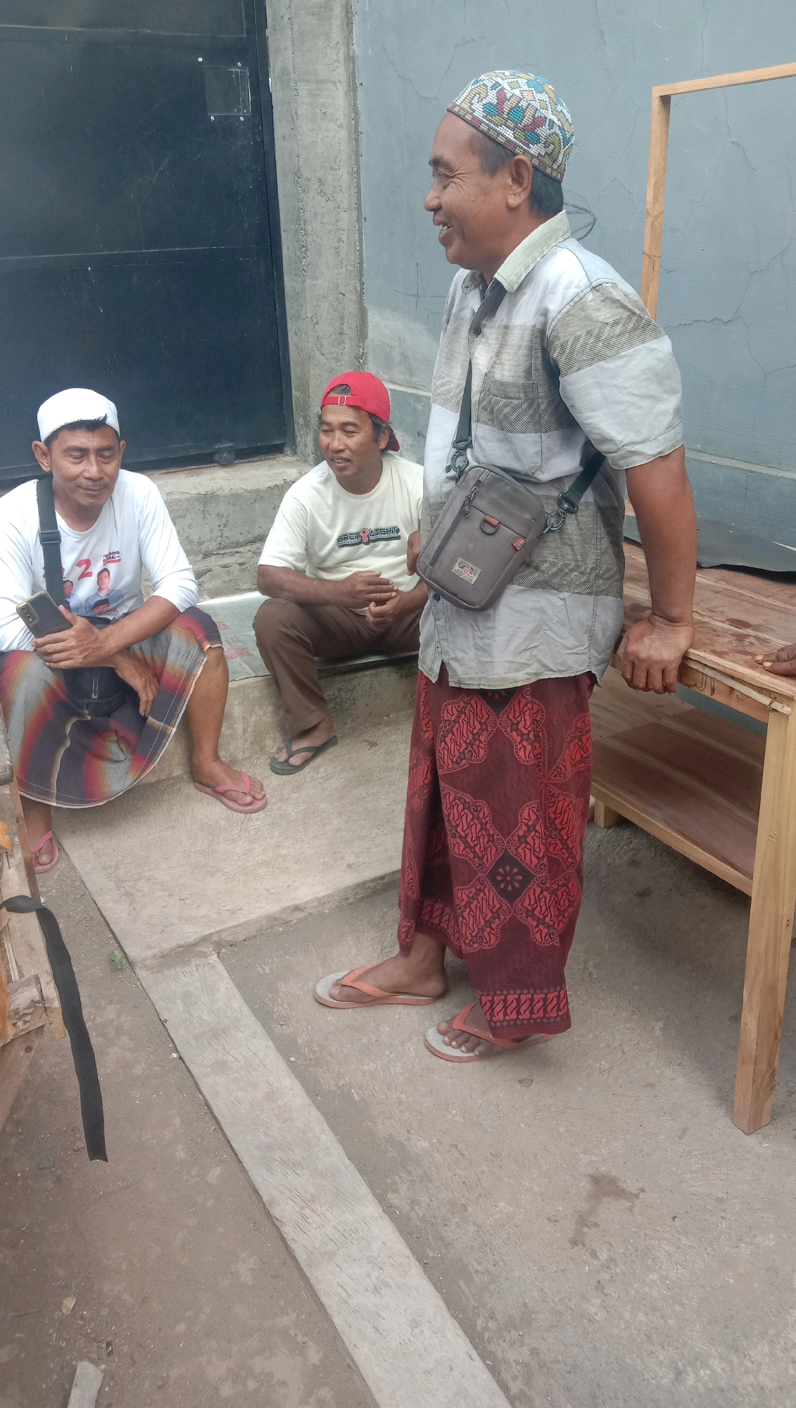 Three men are outdoors in what appears to be a residential area in Karang Anyar, Indonesia. The man on the right, wearing a light-colored patterned shirt, a multi-colored peci hat, and a dark red batik sarong, stands leaning on a wooden table. He has a brown sling bag over his shoulder and is smiling. The man in the middle, wearing a white t-shirt and a red baseball cap, sits on concrete steps. The man on the left, wearing a white t-shirt and a white peci hat, also sits on the steps, holding a black smartphone. He is wearing a sarong with diagonal stripes of red, black, and white. The background features a dark metal door and a grey concrete wall. The ground is made of concrete and dirt.