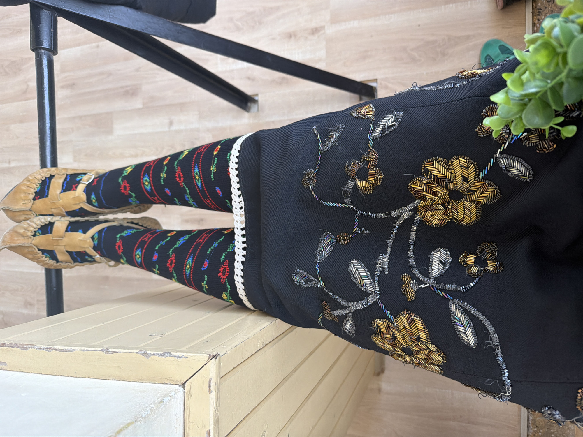 A close-up shot shows the lower half of a person wearing traditional Bulgarian clothing. The person is sitting on a cream-colored, painted wooden chest with horizontal slats. They are wearing black pants or a skirt with ornate gold and silver floral embroidery. The pants are also trimmed with white lace and black fabric featuring colorful patterned socks in red, blue, green, and yellow. Their feet are adorned with tan leather shoes, known as "tservuli". A black metal chair leg is visible in the upper left, and a potted green plant is partially visible on the right side. The background shows a light-colored wooden floor. The location is potentially in Sofia, Bulgaria, due to the traditional Bulgarian clothing.