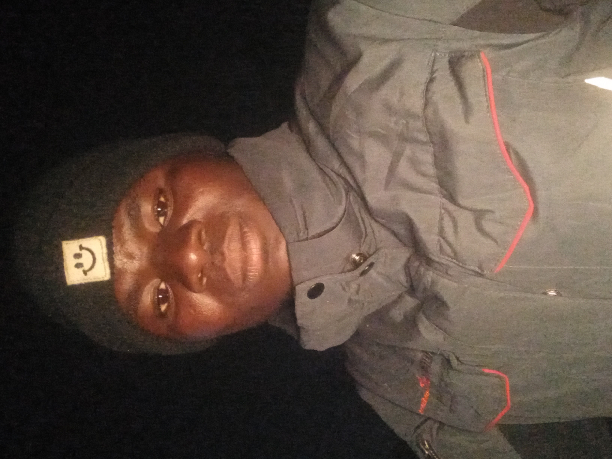 The image shows a close-up, head-and-shoulders shot of a dark-skinned individual wearing a dark beanie with a smiley face emblem and a dark puffer jacket with red trim. The background is entirely black, suggesting a dark environment. The lighting is somewhat dim, casting shadows and highlighting the subject's face. There are no discernible background elements that would indicate a specific location in Kwang, Nigeria, or any other city. The image is a selfie, as indicated by the framing and perspective.