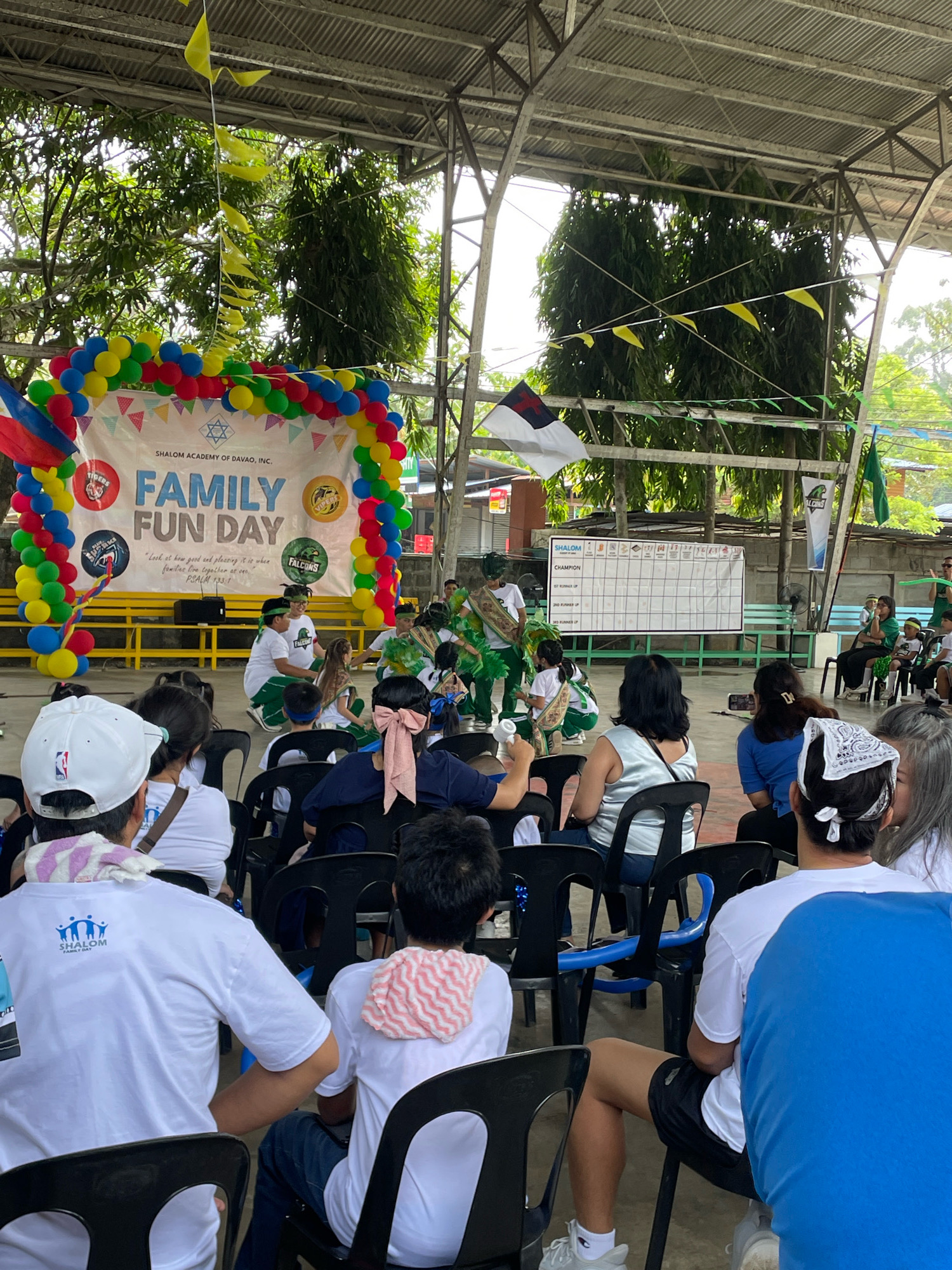 The image depicts an outdoor event held under a covered court, likely at Shalom Academy in Davao City, Philippines, as suggested by the banner. The event, labeled "FAMILY FUN DAY," features a group of children dressed in green and white costumes performing on a stage decorated with a balloon arch and flags. Spectators, including families and students, are seated on black plastic chairs, observing the performance. A scoreboard is visible in the background, indicating a competition or event ranking. The overall atmosphere appears festive and community-oriented.