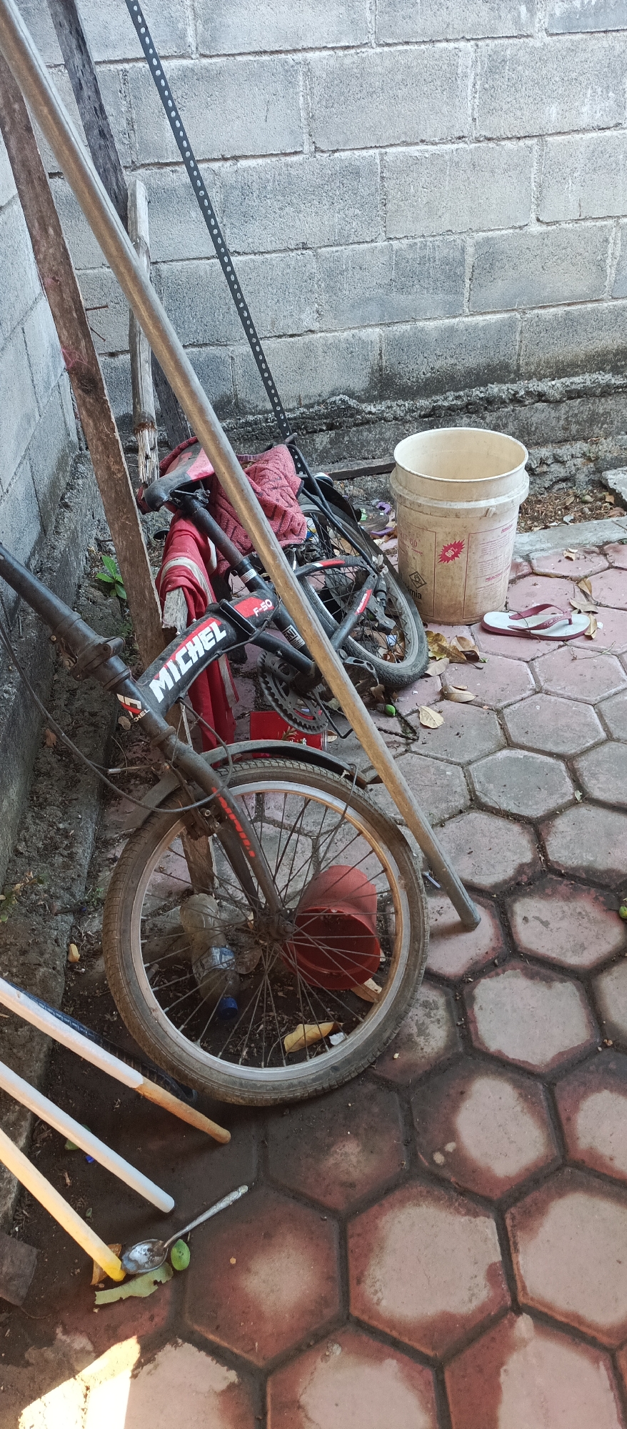 In Arjasa, Indonesia, this image captures a scene outside a residence, likely a yard or alleyway. A broken black bicycle lies on its side against a rough, gray cinder block wall. The bicycle's frame shows the brand name "MICHEL" and "F-50". A red towel or cloth is draped over the bicycle seat and handlebars.
In the foreground, the front wheel of the bicycle is prominently displayed, with a red plastic object and a plastic bottle visible near it. To the left of the bicycle, a tall, thin metal pole leans against a wooden post. A set of white garden tools with wooden handles rests on the hexagonal tiled ground.
To the right of the bicycle, a weathered, off-white plastic bucket stands upright. A pair of pink flip-flops lies on the ground next to the bucket. The ground itself is paved with interlocking hexagonal tiles, some of which appear damp or stained. Scattered leaves and debris are visible on the ground, suggesting an outdoor, potentially untidy, environment. The overall impression is one of disarray and neglect.