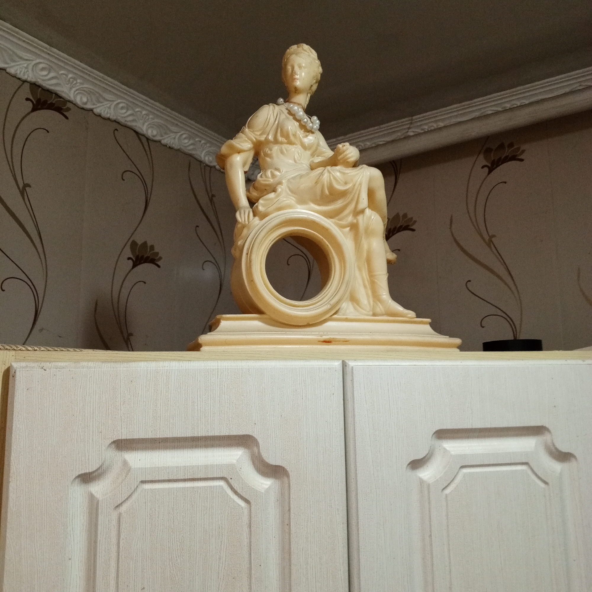 A cream-colored, resin statue of a seated woman with a pearl necklace adorns the top of a light-colored cabinet. The woman is depicted in a classical style, wearing draped clothing. Her body is positioned to the side, with her legs crossed and her hands resting on her lap. A large, circular opening is present in the center of the statue, suggesting it may be part of a clock mechanism. The statue is placed on a shelf above the cabinet doors, which have a subtle wood-grain texture and decorative panels. The background consists of patterned wallpaper with floral motifs and a white, ornate trim along the ceiling line. The overall lighting suggests an indoor setting, likely in a home. The image was taken in Sayanogorsk, Russia.