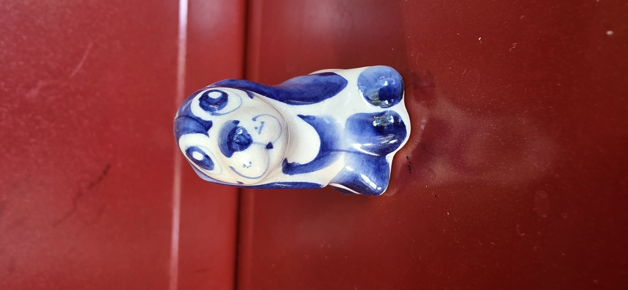 A small, ceramic figurine of a dog, painted with blue and white Gzhel-style patterns, sits on a dark red, glossy surface. The dog figurine is depicted in a sitting pose, with large, expressive eyes and floppy ears. The surface it rests on reflects the figurine and the red background, which appears to be a wall or a piece of furniture. The scene is dimly lit, with the primary light source coming from the front, highlighting the glossy texture of the red surface and casting a soft shadow behind the figurine. The overall impression is one of a simple, decorative object placed on a plain background.