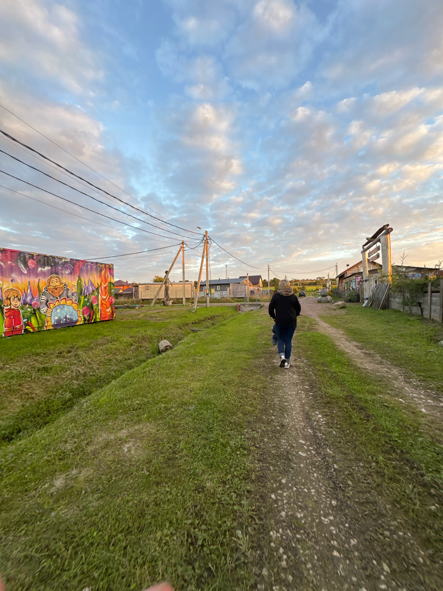 An adult, viewed from behind, walks down a dirt road in Kempelevo, Russia, towards a cluster of houses and outbuildings. The individual wears a dark hooded jacket, blue jeans, and dark sneakers with white soles. A large, colorful metal container adorned with graffiti art stands to the left of the road. Overhead, power lines stretch across the sky, which is filled with a mix of blue sky and fluffy white and pink clouds, suggesting a late afternoon or early evening setting. The road is bordered by green grass, and a small ditch is visible to the left. In the distance, more houses, fences, and vegetation indicate a rural or semi-rural settlement. The overall scene depicts a quiet, casual moment in a village environment.