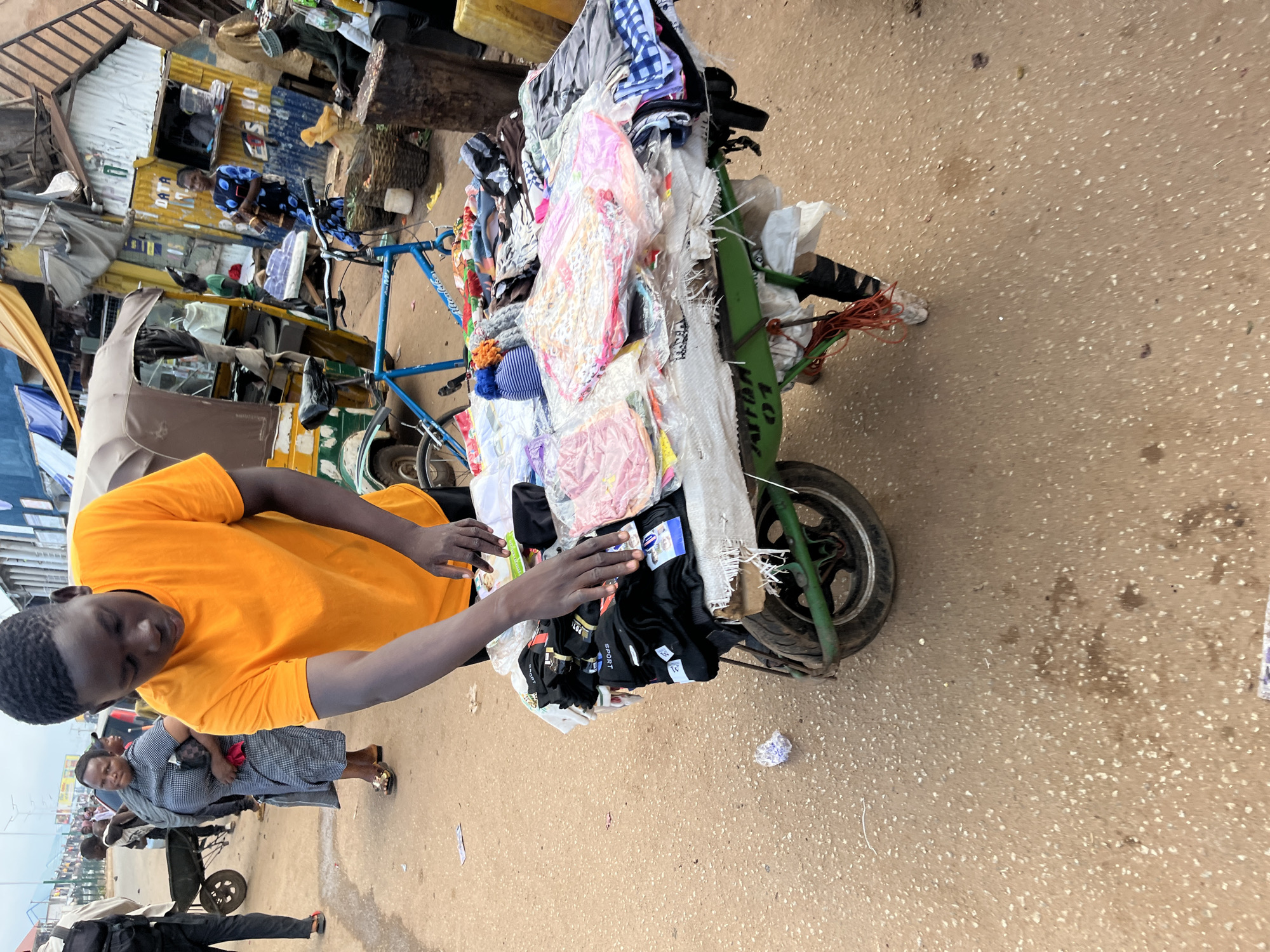 A young man in an orange t-shirt is standing behind a cart filled with clothing, likely at a market stall in Bukuru, Nigeria. He is reaching out to the clothing, appearing to be sorting or displaying it. The cart is old and appears to be made from repurposed materials, with a green frame and a single wheel. Various items of clothing are spread across the cart, some neatly folded and others in plastic packaging. In the background, other people are visible, as well as market stalls and a three-wheeled vehicle. The ground is a sandy, dirt surface, typical of an outdoor market. The overall scene depicts a busy commercial environment.