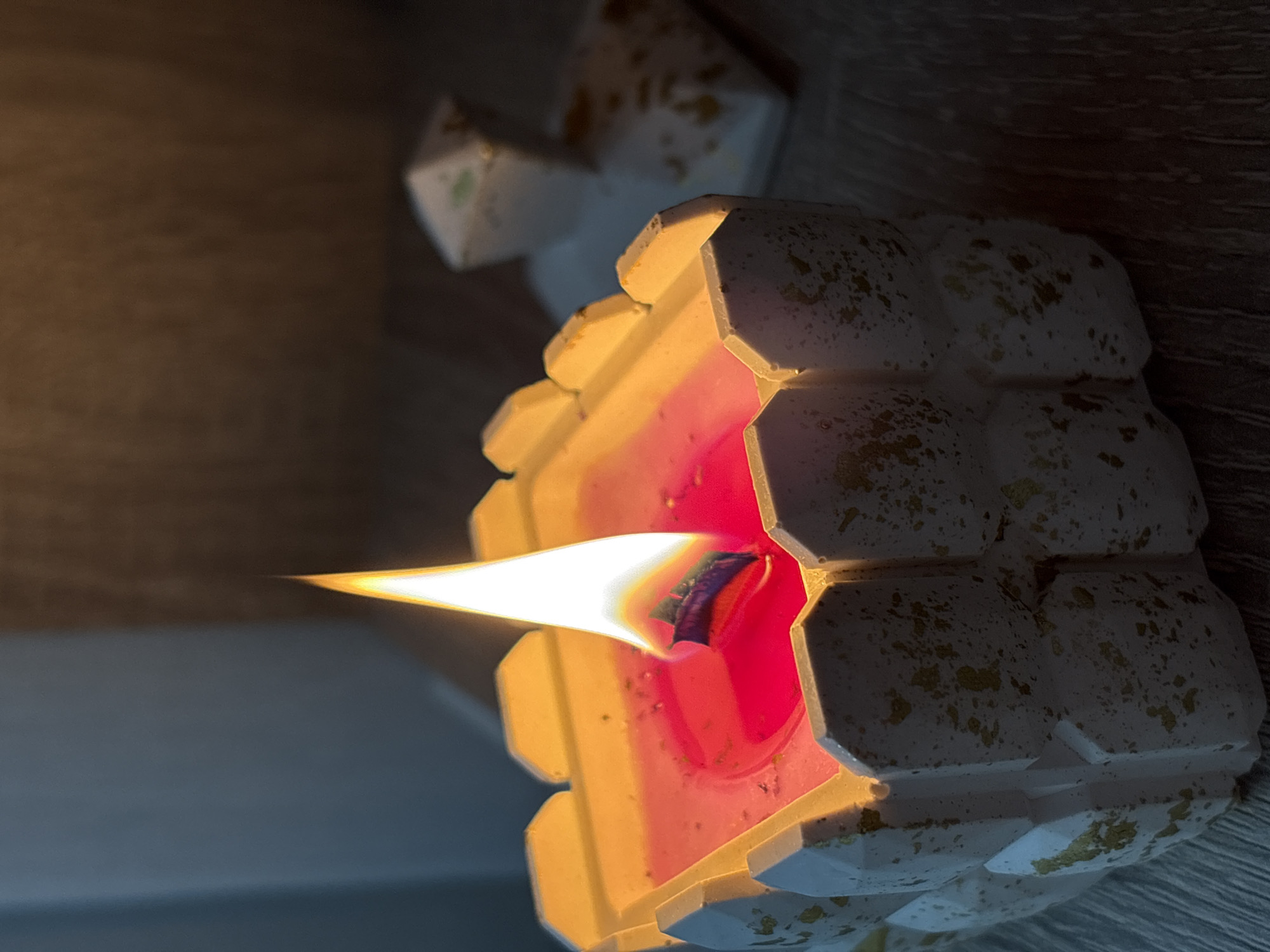 A lit candle sits on a wooden surface. The candle's outer casing is a geometric white with gold specks. The candle wax inside is red, with the flame extending outward. The background is a combination of light and dark wood. Another white geometric object is behind the candle. The location is not known to be Birobidzhan, Russia.