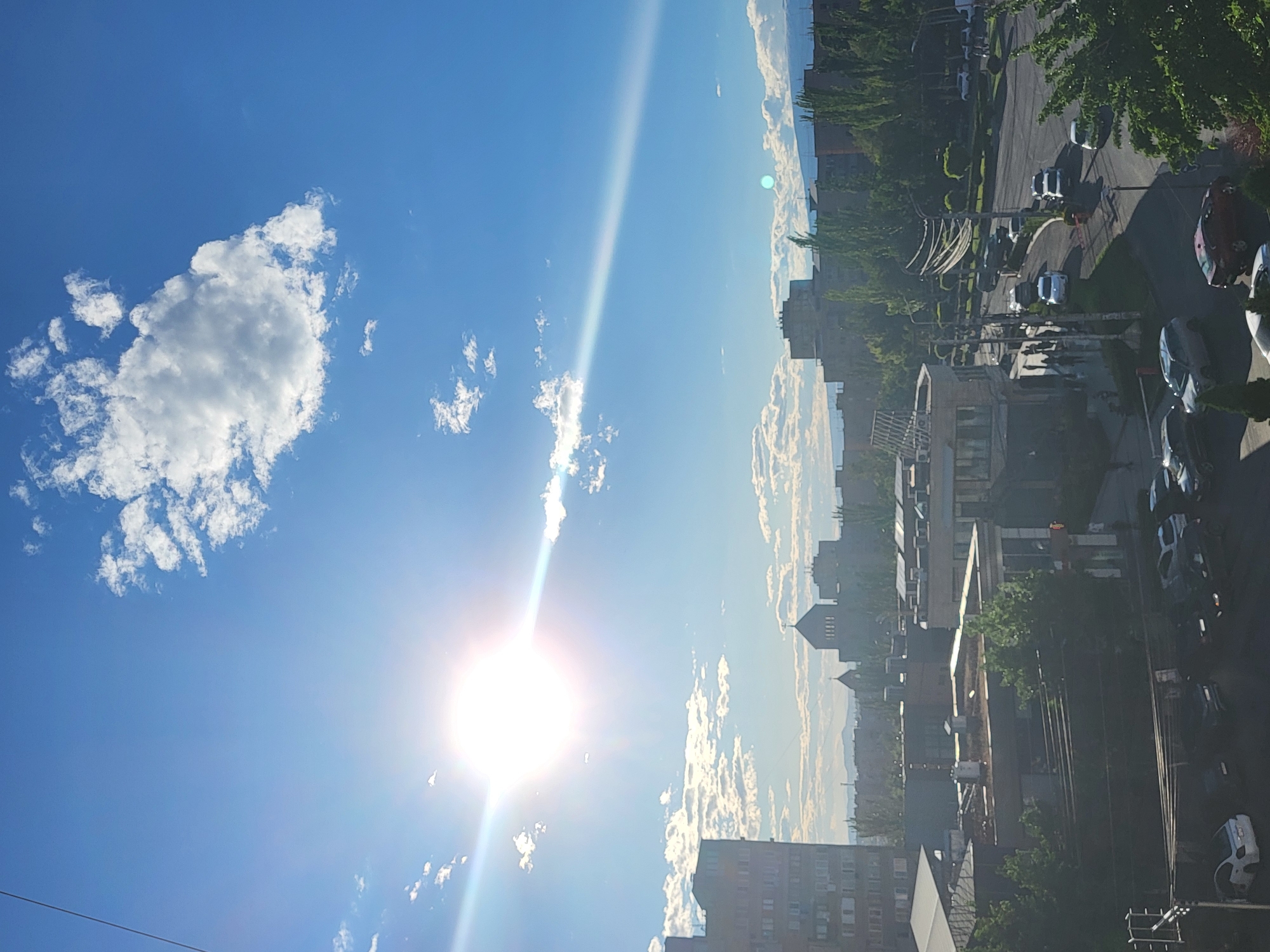 The image presents a daytime cityscape under a bright sky, potentially taken from a high vantage point. The sky, which takes up the majority of the frame, is a clear blue with scattered white clouds. The sun is prominently positioned, emitting a strong glare. Below the sky, there's a view of a city with buildings of varying heights, suggesting a densely populated area. Roads lined with parked cars and interspersed with trees are visible. Specific identification of the city as Yerevan, Armenia is not possible from the visual information.