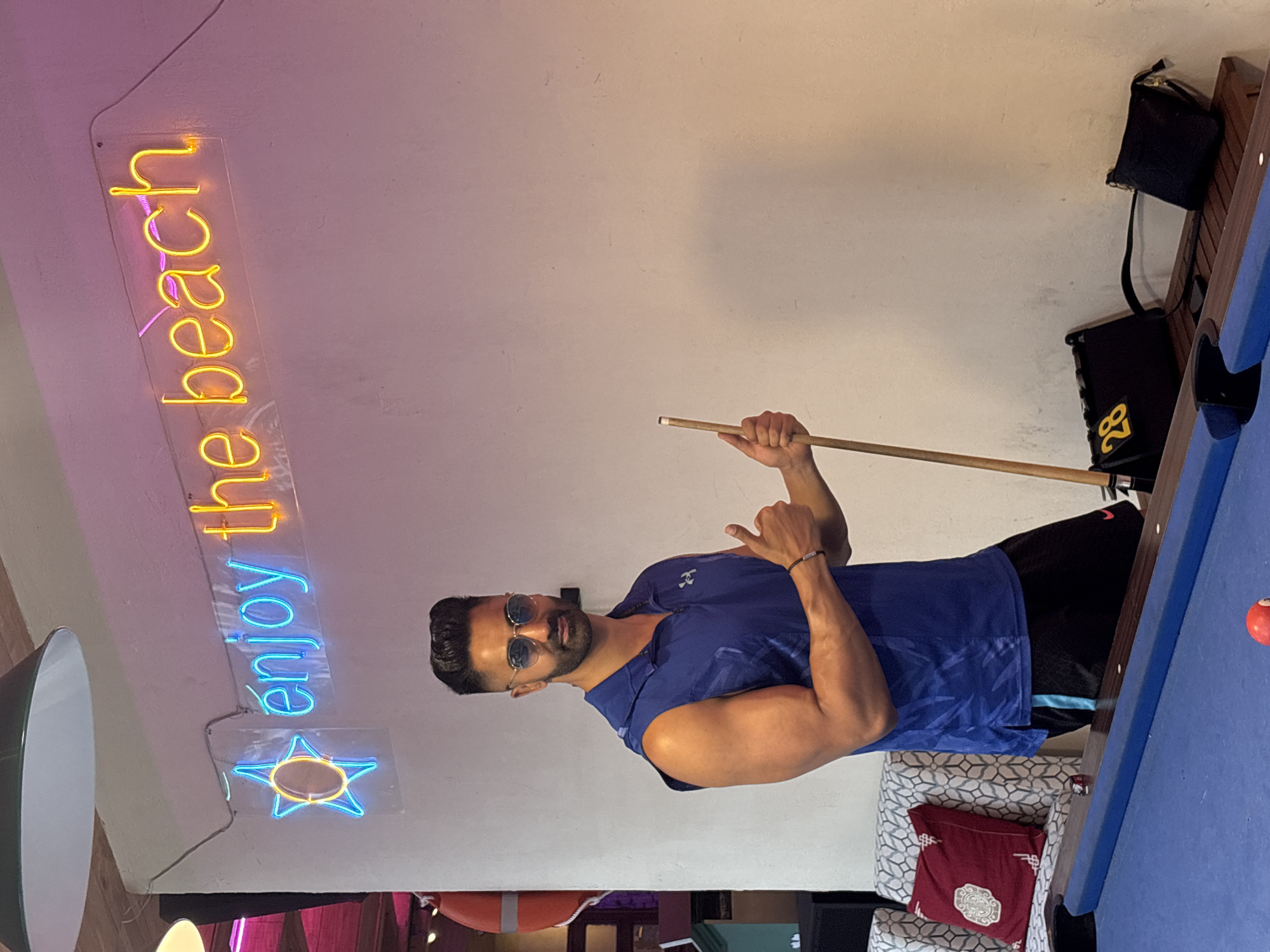 A man stands next to a pool table in an indoor setting. He wears a blue sleeveless shirt, black shorts, and sunglasses. He holds a pool cue in his right hand, pointing his left index finger upward. In the background, a neon sign reads "enjoy the beach" above a neon star. Behind the pool table, there is a patterned chair and bags hanging on the wall. The pool table is blue, with a visible pool ball on the surface. Given the neon sign and the overall setting, the image is potentially taken in a recreational or entertainment venue, possibly in a tourist area. Without additional information, it is impossible to confirm the exact location within Đà Nẵng, Vietnam.