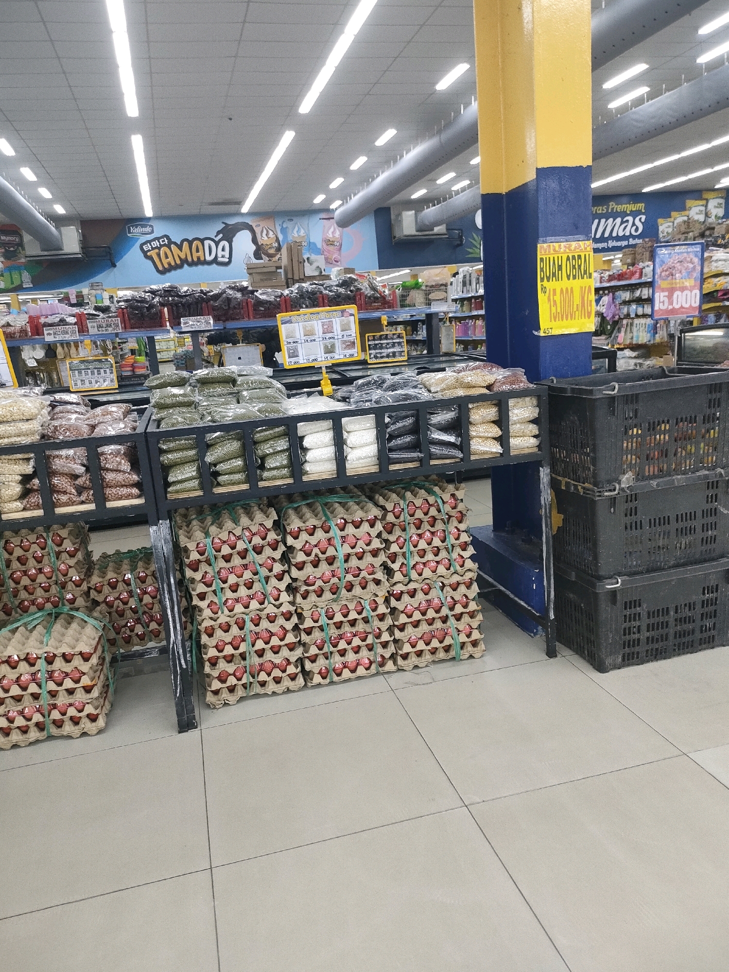 The image shows a retail store interior, likely a supermarket, in Batam, Indonesia. The scene is dominated by shelves stocked with various food items. In the foreground, stacks of egg cartons are visible, some tied with green twine. To the right, large black plastic crates are stacked. Behind the egg cartons, shelves display bags of dried goods such as beans and grains. A yellow and blue pillar, likely part of the store's structure, is prominent on the right side, with a yellow sign attached that reads "BUAH OBRAL" (fruit sale) and pricing information. In the background, more shelves with products are visible, along with store lighting and ceiling fixtures. A large banner with the text "Yalindo" and "TAMADA" is displayed on the back wall, suggesting a brand or store name. The overall environment appears clean and organized, typical of a commercial grocery setting.
