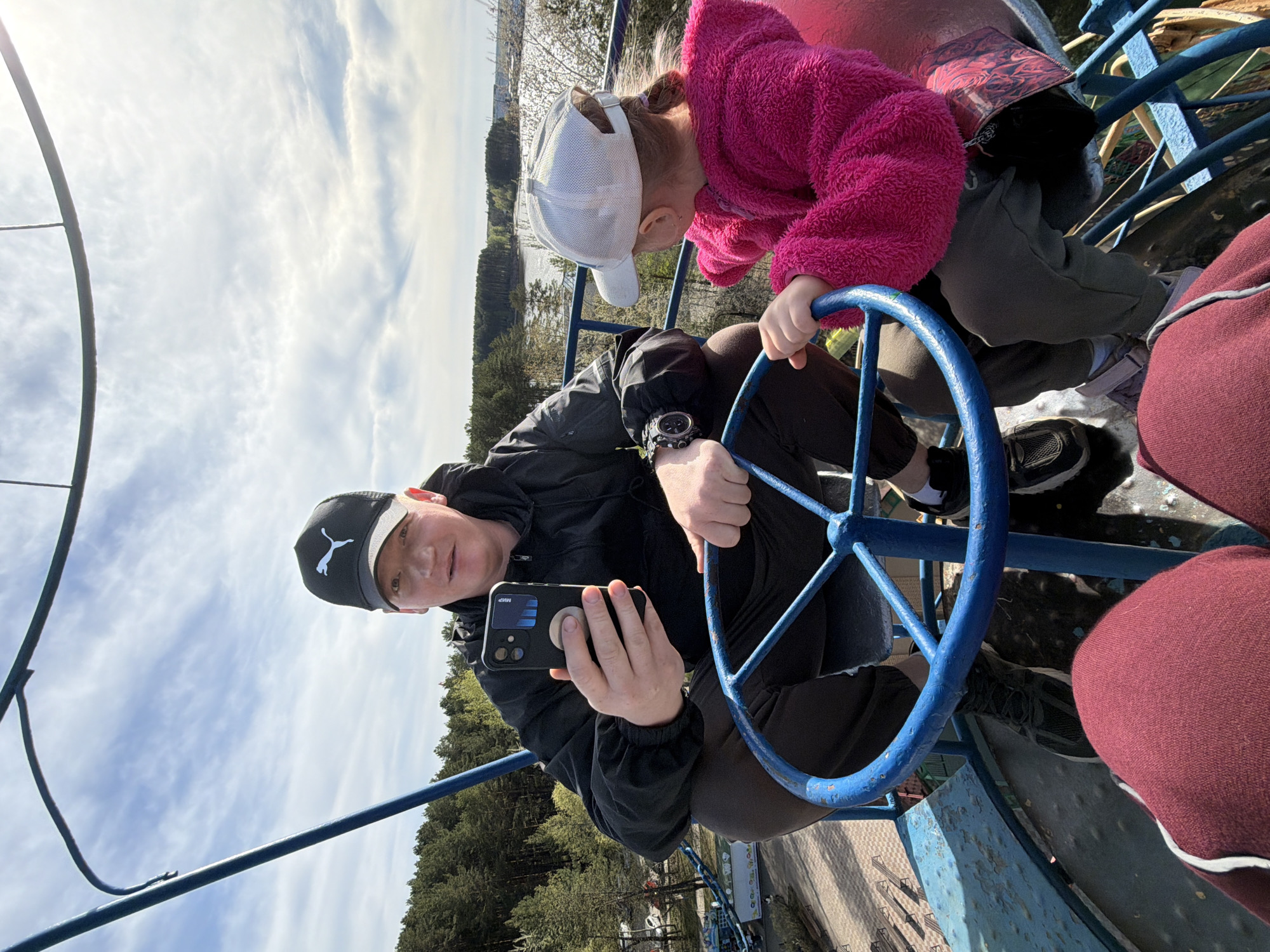 A man and a child are seated on a structure with a blue steering wheel, possibly a small carousel, located in Chaikovsky, Russia. The man, wearing a black Puma cap, a black jacket, and a watch, is holding a black smartphone and looking at the camera. A girl, in a white cap and pink jacket, is looking down at something below. The background shows a sky with clouds, trees, and a view of a body of water in the distance.