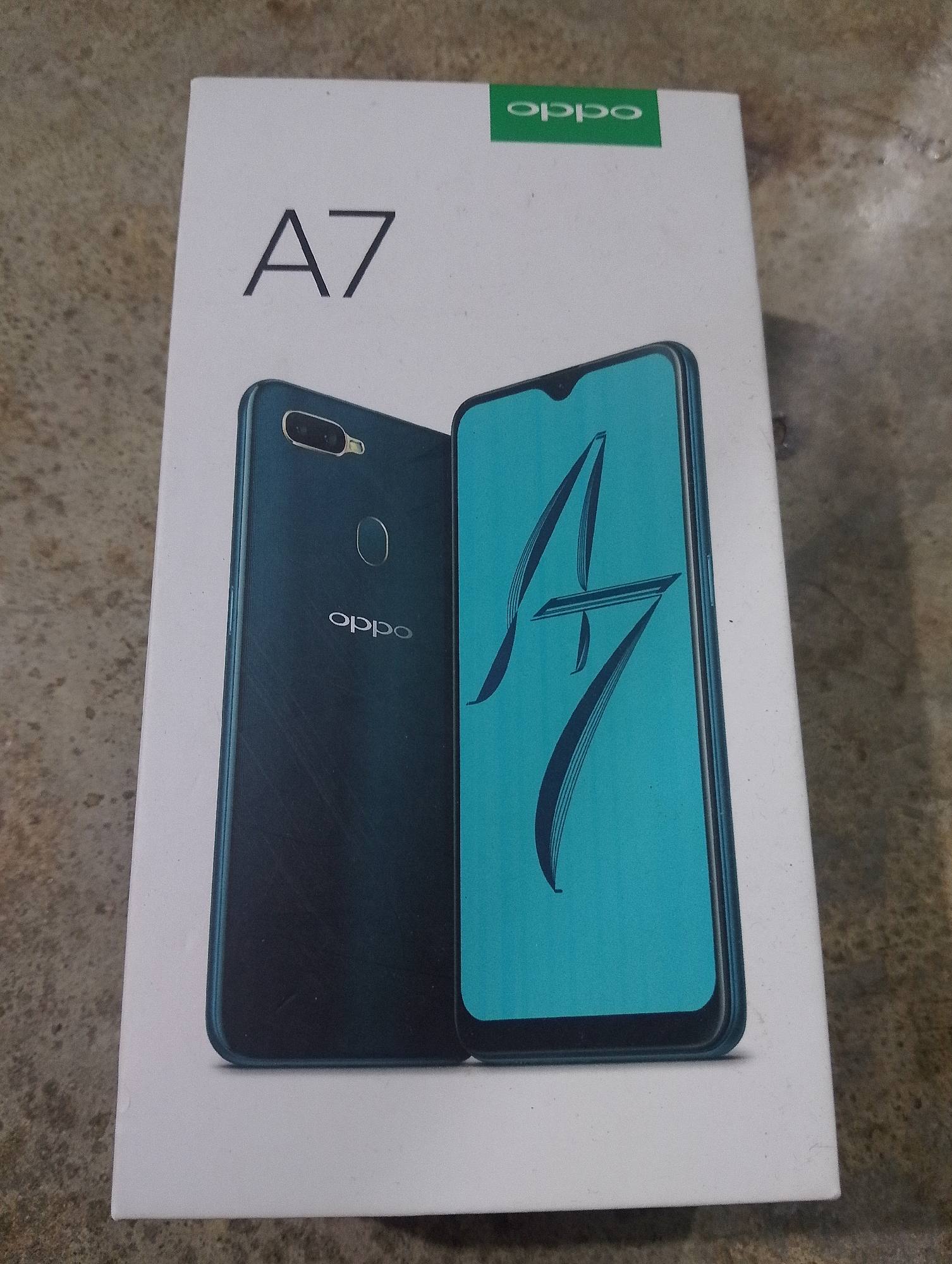 The image shows the retail packaging of an Oppo A7 smartphone. The box is white with the "A7" model name prominently displayed in black text. A green Oppo logo is visible in the top right corner of the box. Two images of the Oppo A7 phone are depicted on the box: one showing the rear of the phone with a dual-camera setup and the Oppo logo, and another showing the front of the phone with its teal-colored screen displaying the "A7" logo. The box rests on a textured surface that appears to be concrete or stone. The overall scene is a static product shot. There are no people or any indication of the city of Serbelawan, Indonesia.