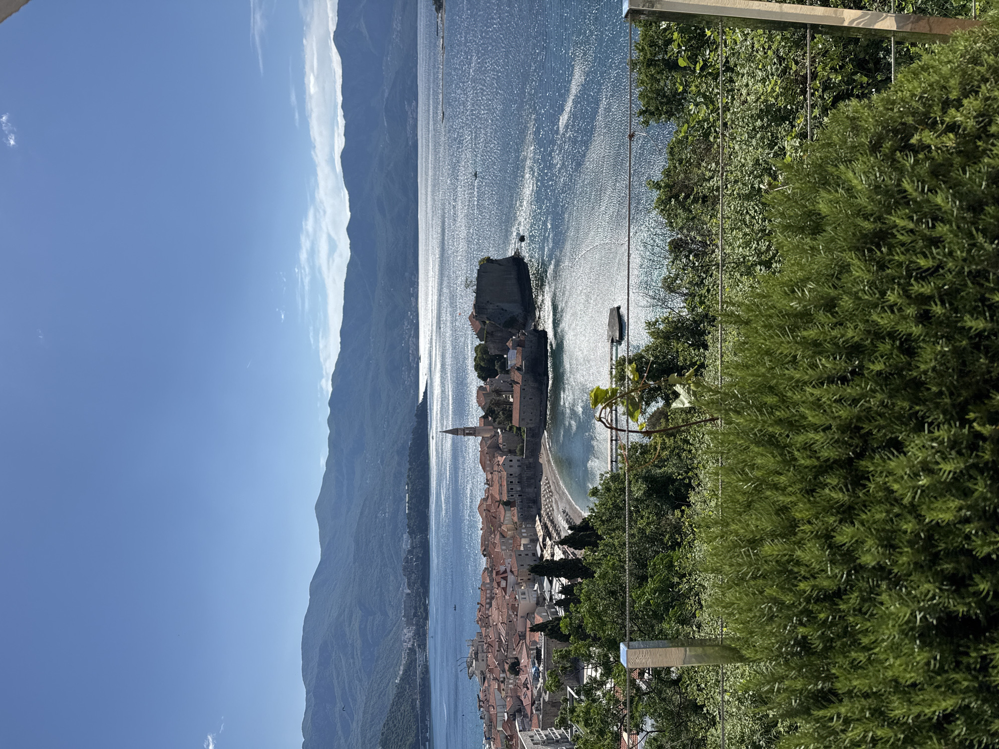 The image shows the old town of Budva, Montenegro, viewed from an elevated perspective, possibly from a balcony or terrace. The old town is situated on a peninsula surrounded by the Adriatic Sea, with mountains visible in the background on the mainland. The sea is sparkling with sunlight. The old town's characteristic red-tiled roofs and a prominent church tower are visible. The foreground features greenery, including bushes and trees, along with a portion of a metal railing or structure.