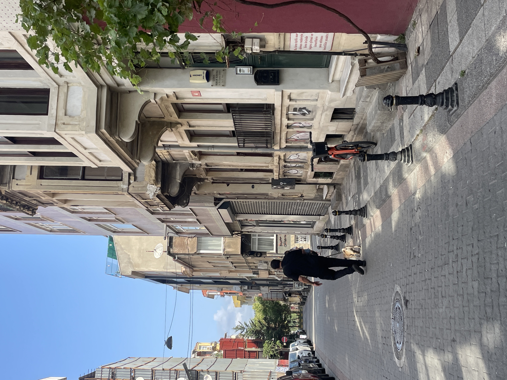 The image depicts a street scene in Kadıköy, Turkey, with a man walking a dog. The man, dressed in a black t-shirt and dark pants, is in the foreground, walking away from the camera on a cobblestone street. He is holding a leash attached to a small, light-colored dog that is walking beside him.
The street is lined with buildings of varying architectural styles. On the right side of the street, a building features ornate detailing, including a bay window with decorative stonework and a wrought-iron railing. A red vine climbs the facade of this building. A bright orange bicycle is parked near the building. A sign with Turkish text is visible on the building's exterior.
On the left side of the street, the buildings appear more modern, with some showing signs of scaffolding or construction. Several cars are parked along the curb.
The sky is clear and blue, indicating a sunny day. The overall impression is of a residential or commercial street in an urban environment. The sunlight casts shadows across the cobblestone pavement, highlighting the texture of the street surface. The scene captures a moment of everyday life in the city.