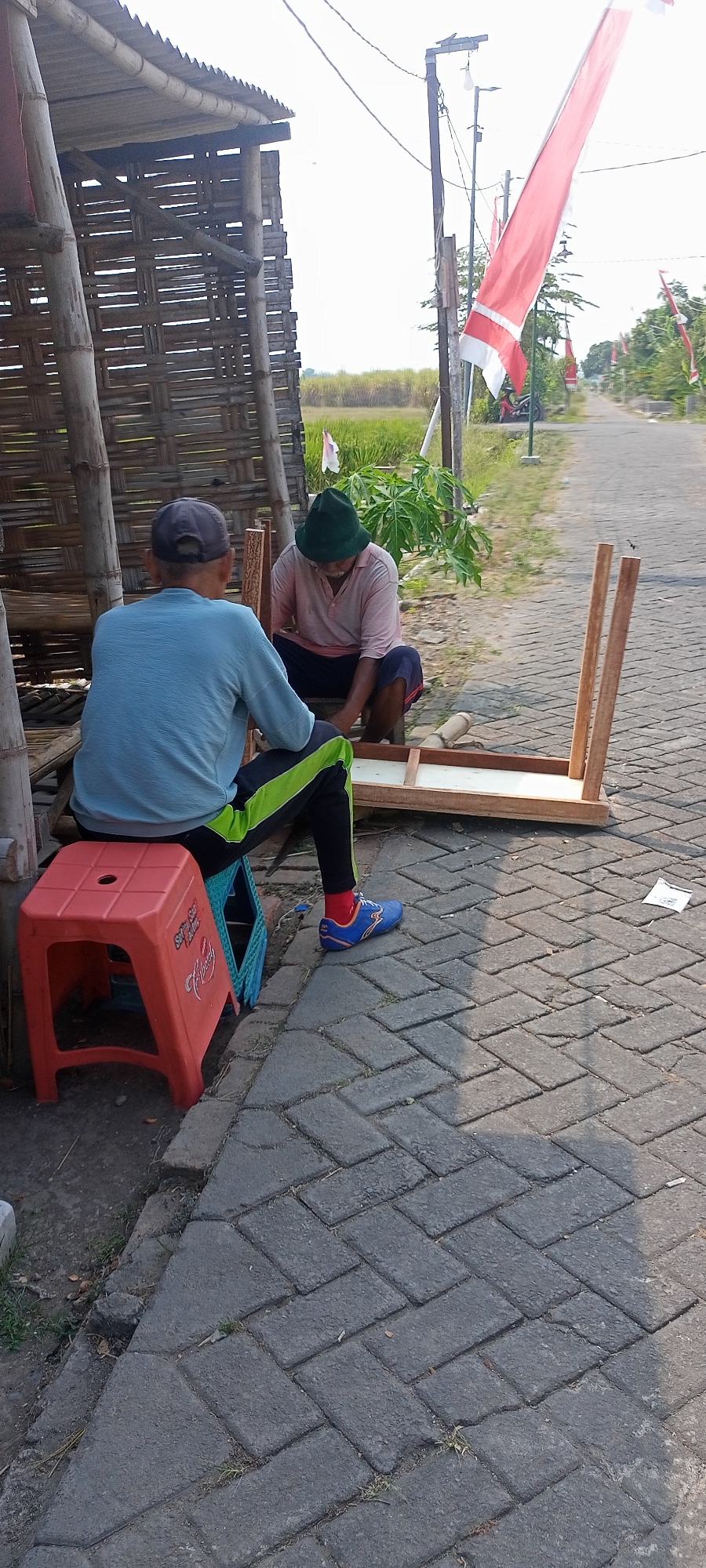 In Gempolklutuk, Indonesia, two men are observed outdoors near a humble, woven bamboo structure. The man on the left, wearing a light blue long-sleeved shirt, dark pants with lime green stripes, a gray baseball cap, and blue athletic shoes with red socks, is seated on a red plastic stool. The man on the right, dressed in a pale pink long-sleeved shirt, dark shorts, and a dark green hat, is kneeling. Between them lies a partially assembled wooden table with two legs attached to a rectangular frame. The ground is paved with interlocking gray bricks. In the background, a paved road extends into the distance, flanked by greenery and several Indonesian flags displayed on poles. A red motorcycle is parked on the side of the road. The overall scene suggests an outdoor workspace or a moment of communal activity.