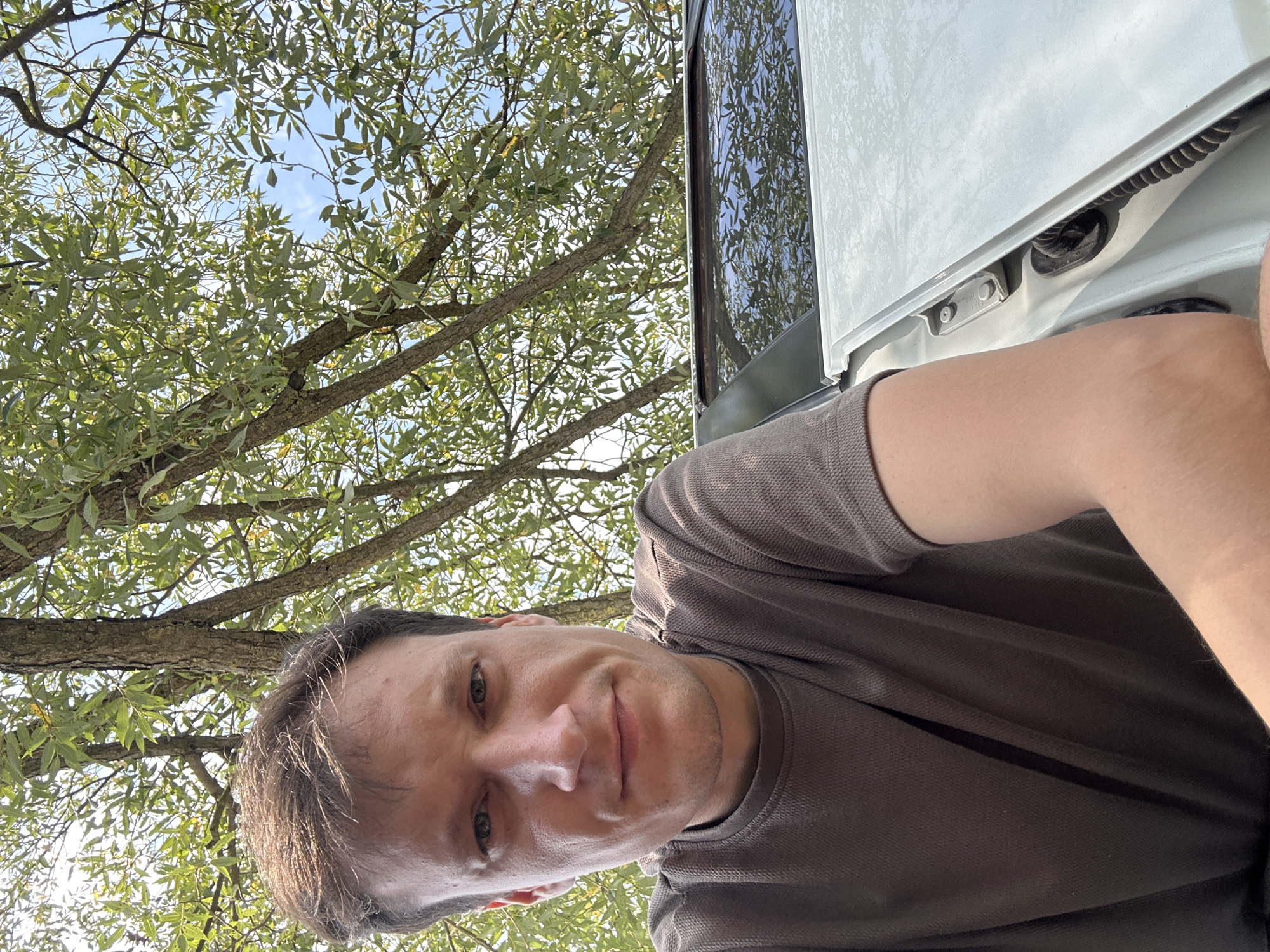 A low-angle medium shot captures a man taking a selfie outdoors, with a bright sky and leafy trees forming the background. The man, who appears to be in his late 30s or early 40s, has fair skin, short brown hair, and is wearing a dark grey, short-sleeved t-shirt. He is positioned in the foreground, slightly to the left of the frame, with his head tilted and a neutral expression. To the right, the hood and windshield of a light-colored vehicle are visible, reflecting the surrounding trees and sky. The environment suggests a park or natural setting within the city of Makiv, Ukraine.
