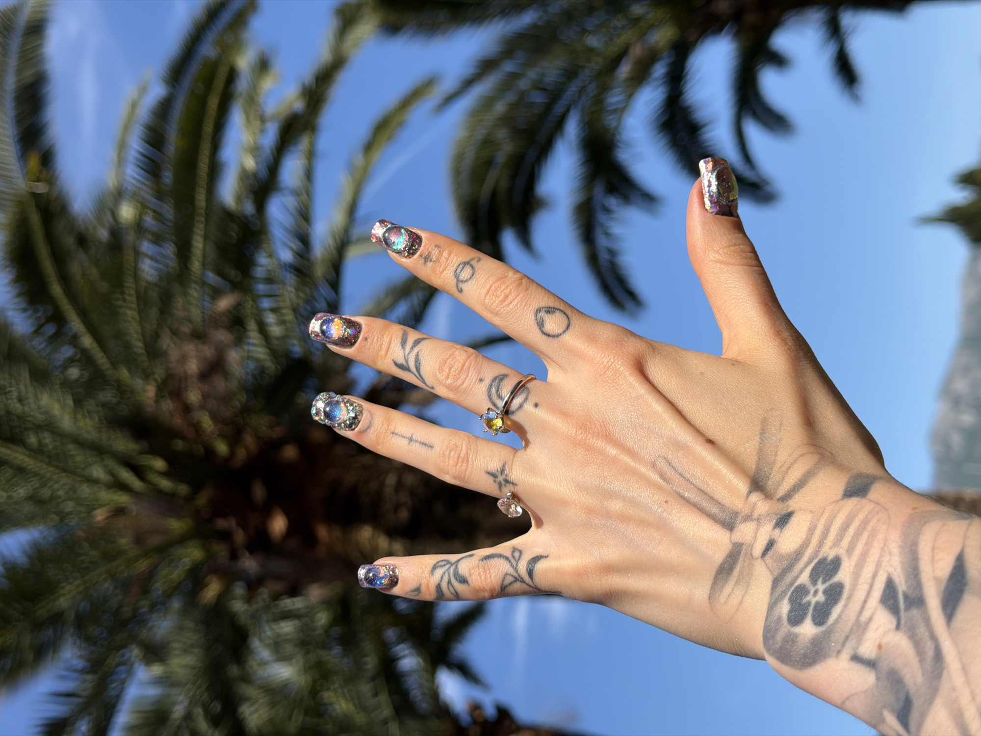 A left hand, adorned with elaborate nail art, rings, and tattoos, is prominently displayed against a vibrant blue sky and blurred green palm fronds. The hand is held up, showcasing its unique features in bright daylight.
The fingernails on all visible fingers feature a dazzling, iridescent galaxy or cosmic-themed design, incorporating glitter, specks of white, and swirls of blues, purples, and golds that catch the light.
The fingers are decorated with several small, minimalist stick-and-poke style tattoos, including abstract shapes, a crescent moon, and a small circle. On the middle finger, a delicate gold-toned ring with a small, faceted, multi-colored stone is worn. Another similar ring, possibly with a clear or light-colored stone, is visible on the ring finger.
Extending from the base of the fingers across the back of the hand and onto the wrist/lower forearm are more intricate and larger tattoos. These feature flowing, organic lines that resemble floral or leafy patterns, blending into a more stylized, abstract design that appears to incorporate elements of a complex insect or a geometric motif near the wrist.
The background consists of out-of-focus, dark green palm leaves, suggesting a warm, possibly coastal environment, consistent with Budva, Montenegro. The sky is a clear, bright azure, indicating a sunny day. No discernible text is visible in the image.