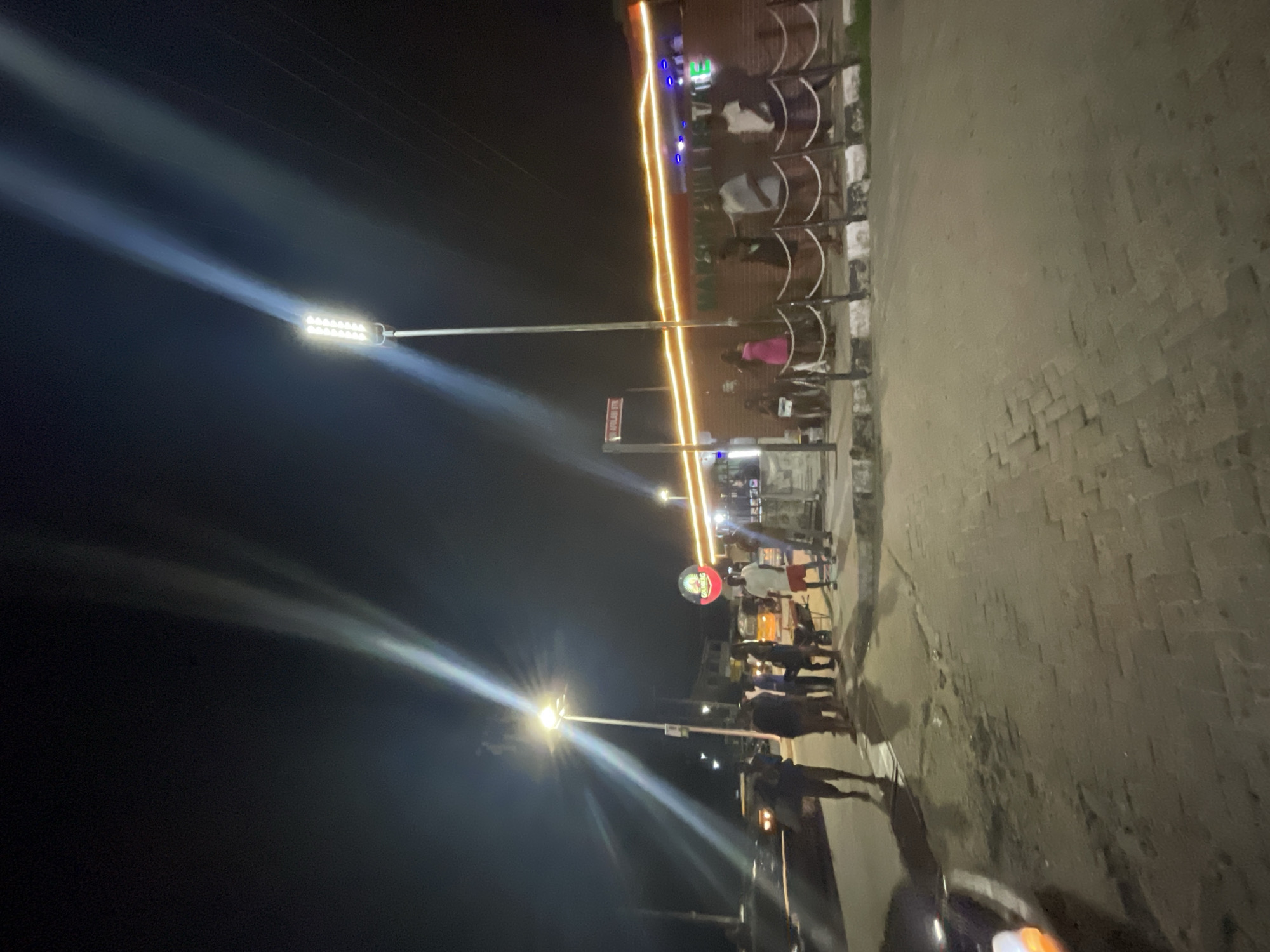 This nighttime image captures a lively street scene, likely in Ikorodu, Nigeria, centered around an outdoor establishment. Two prominent street lights, equipped with multiple LED panels, illuminate the area with bright, distinct beams cutting through the dark sky.
Several people are gathered along the right side of the frame, either seated on stools with curved backrests or standing. They appear to be patrons or workers, socializing or engaged in activities at the establishment. Their faces are not clearly visible due to the low light and distance.
The building behind the people is adorned with warm, horizontal strip lights, suggesting a commercial space like a restaurant, bar, or shop. Above the crowd, a circular sign with a white background and red text clearly displays the word "GOODIES." Below the building's main lighting, a rectangular sign is partially visible, with faint text that appears to read "HAPPINESS ATE" or "HAPPINESS HERE."
The ground is paved, showing textures consistent with interlocking bricks or concrete. In the bottom right corner, the front section of a vehicle is visible, distinguished by an illuminated orange indicator light. The overall setting suggests a bustling night environment with a social atmosphere. The sky is dark, confirming it is nighttime, and the absence of weather phenomena indicates clear conditions.