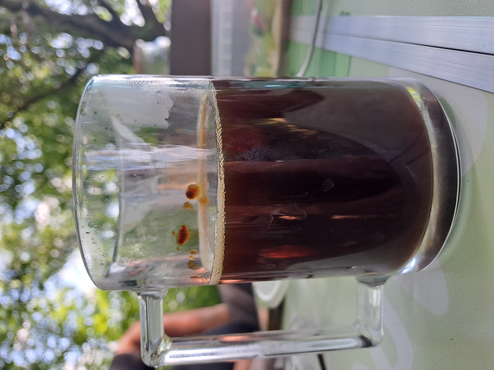 A glass mug filled with dark liquid, likely coffee, sits on a light green patterned surface. The mug has a thick handle and the glass is slightly smudged with water droplets and some small dark particles visible on the inside. The liquid has a thin layer of foam at the top. In the blurred background, foliage and trees are visible, suggesting an outdoor setting. To the right of the mug, two metallic strips are placed parallel to each other. The overall scene appears to be a casual setting, possibly in Dedinovo, Russia, during daylight.
