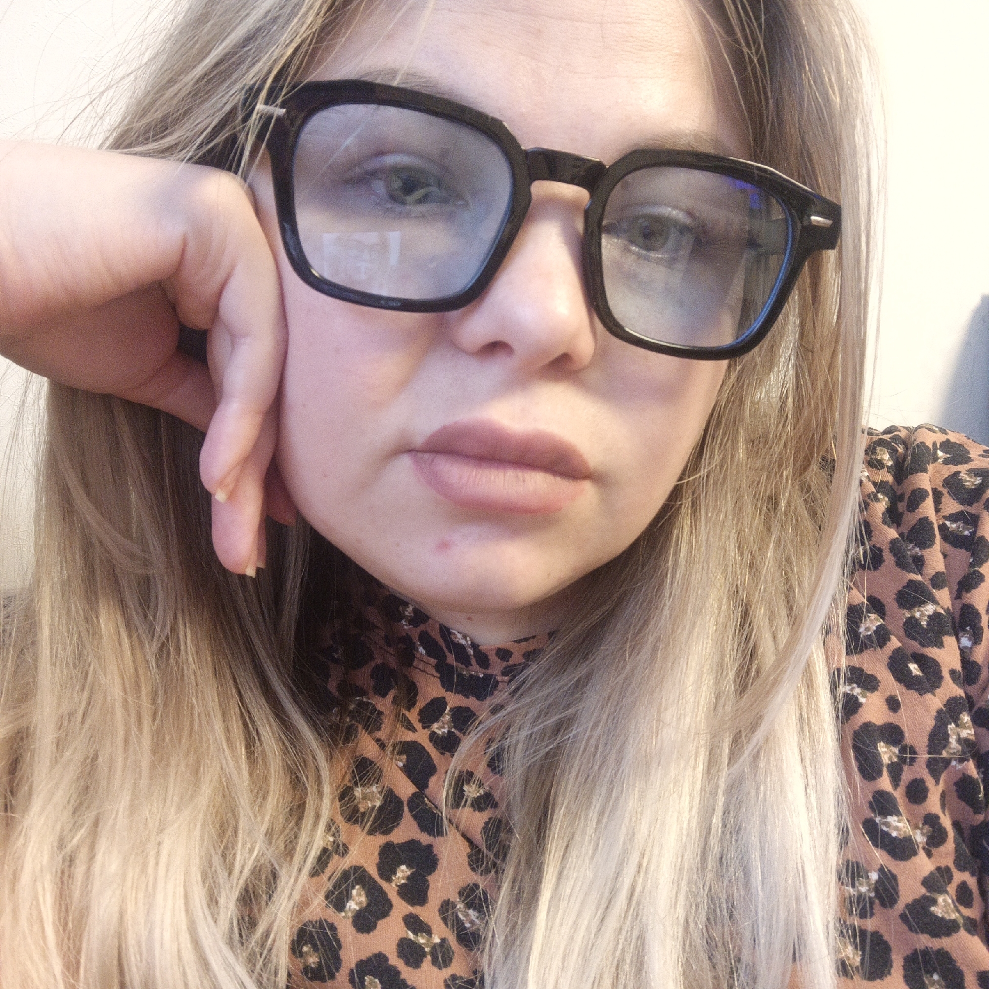 A close-up, head-and-shoulders portrait shows a young woman with long, blonde or light brown hair that falls around her face and shoulders. She is wearing black, thick-framed square glasses through which her light-colored eyes are visible. Her lips are natural or have a light, matte lipstick, and are slightly parted. Her right hand is resting against her face, with her fingers supporting her chin and cheek. A small blemish is visible on her chin. She is wearing a top with a brown and black leopard print pattern, visible on her shoulder and chest area. The background is a plain, light-colored wall, out of focus. The lighting appears soft and even, suggesting an indoor setting. There is no visible text. The woman's expression is neutral and direct towards the viewer.