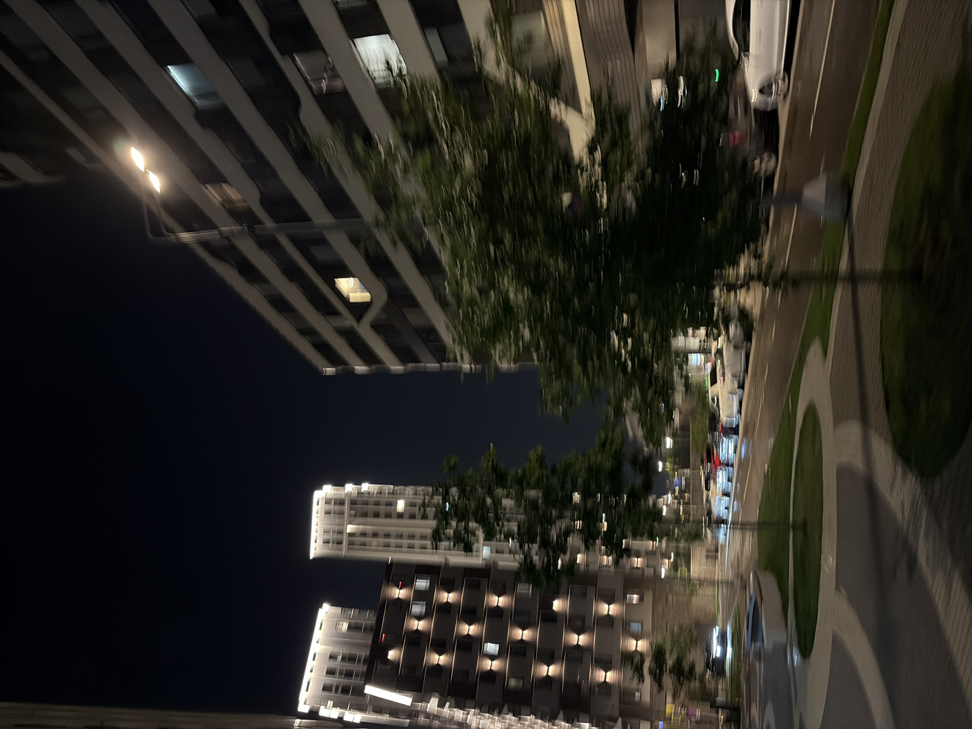 This is a nighttime photograph of a modern urban environment in Moscow, Russia. The scene features several tall, contemporary apartment buildings with illuminated windows, indicating residential occupancy. In the foreground, a street or promenade area is visible, paved with light-colored tiles and bordered by green lawns and trees. There are several cars parked or driving on the street, some of which are white. A street lamp with two glowing lights illuminates the scene. The overall atmosphere is one of a developed, well-lit urban neighborhood at night.