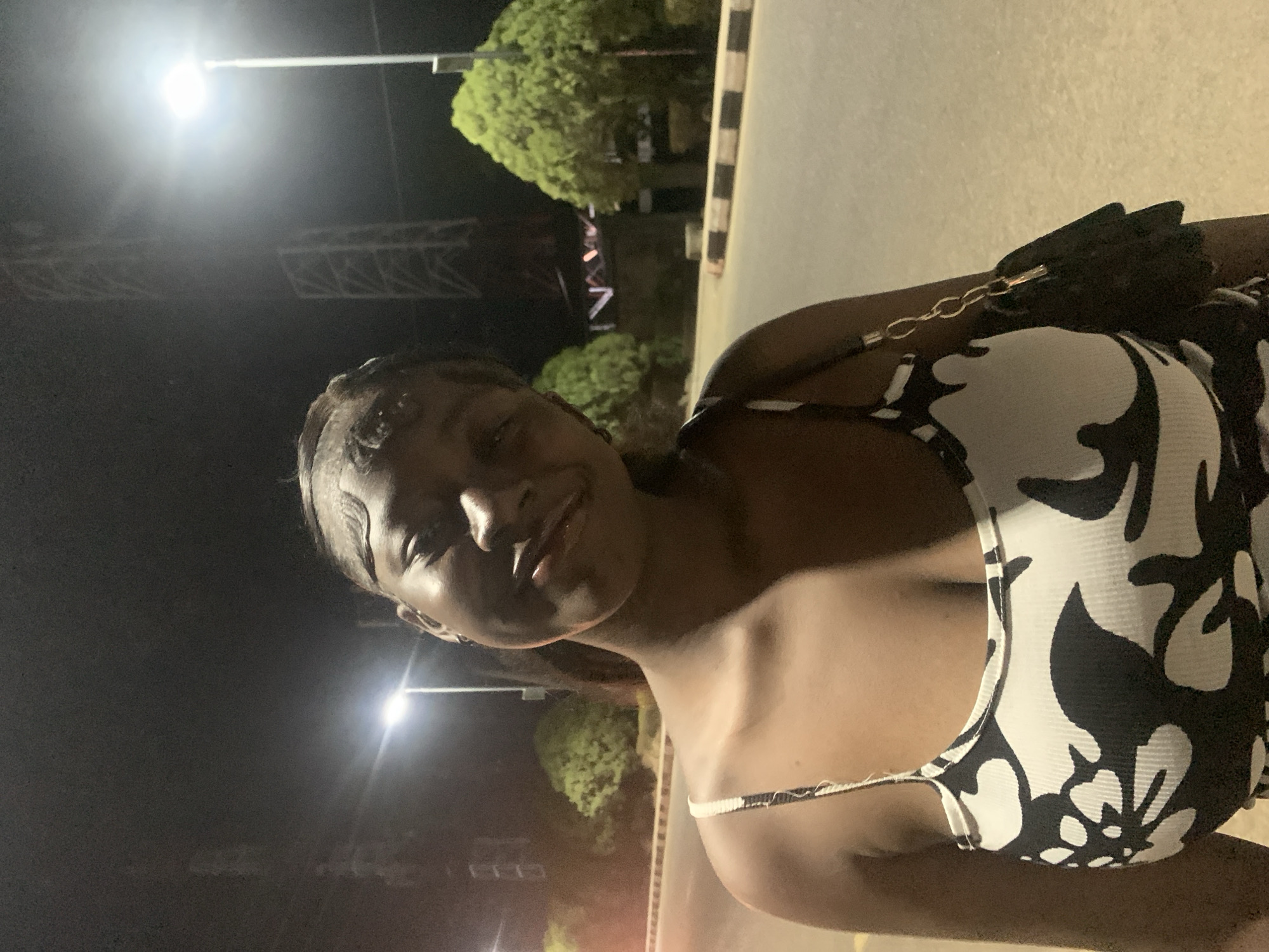 A woman is posing for a selfie at night. She has dark skin and is wearing a white and black patterned top with thin straps. She also has a black purse with a chain strap. Behind her is a street with streetlights and some trees. In the background, there are some structures resembling scaffolding or decorative lighting. The location is likely in Jos, Nigeria.
