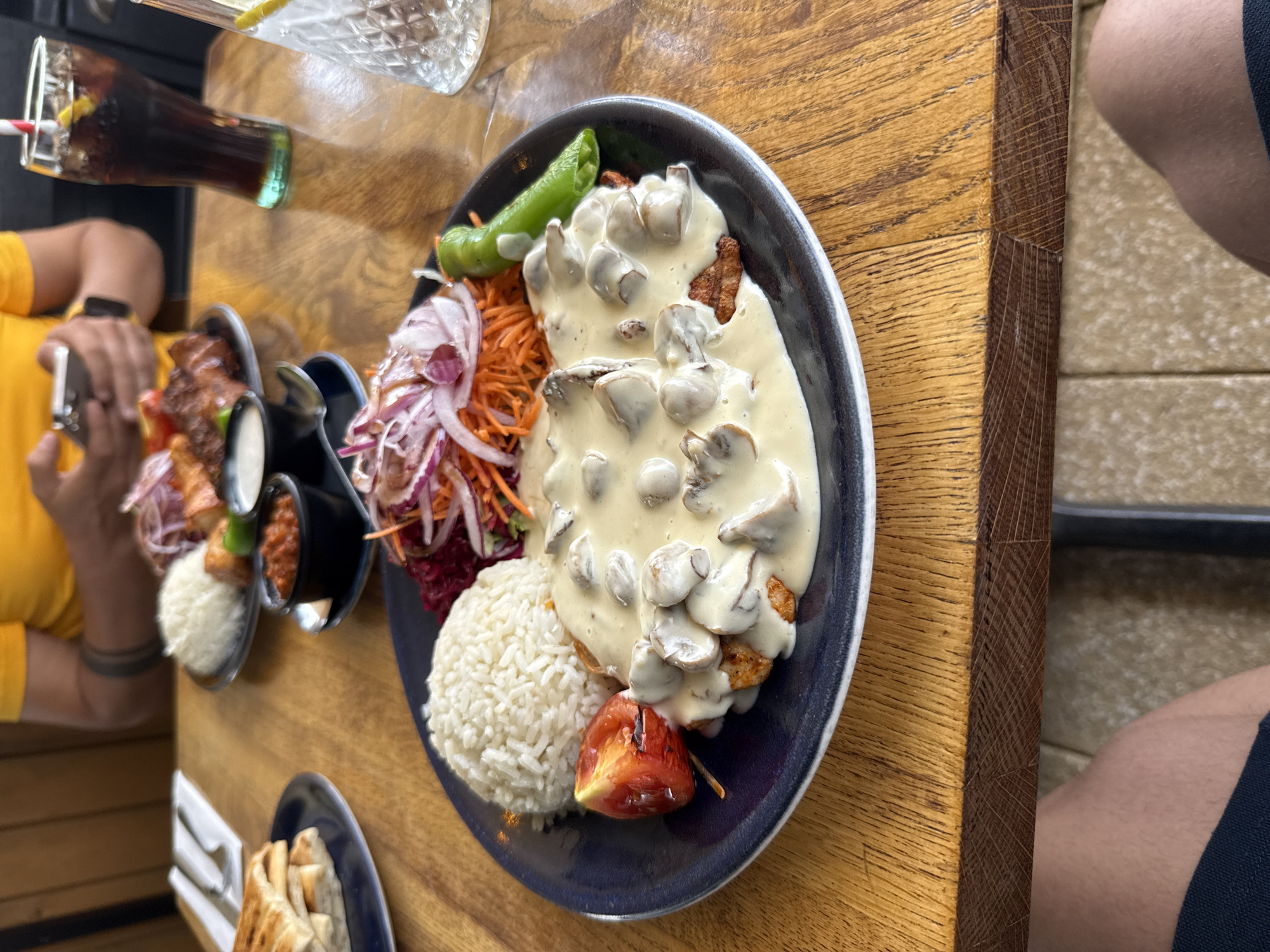 A close-up, overhead shot shows a meal on a wooden table in Braintree, United Kingdom. The meal includes a plate with white rice, grilled meat topped with a creamy mushroom sauce, a grilled tomato, and a salad of shredded carrots and red onions. A green pepper sits on the side of the plate. In the background, another plate of food, a glass of dark soda with a straw, and a glass of water with ice and lemon are visible. A person wearing a yellow t-shirt is partially visible in the background, holding a mobile phone. The table has a visible wood grain texture.