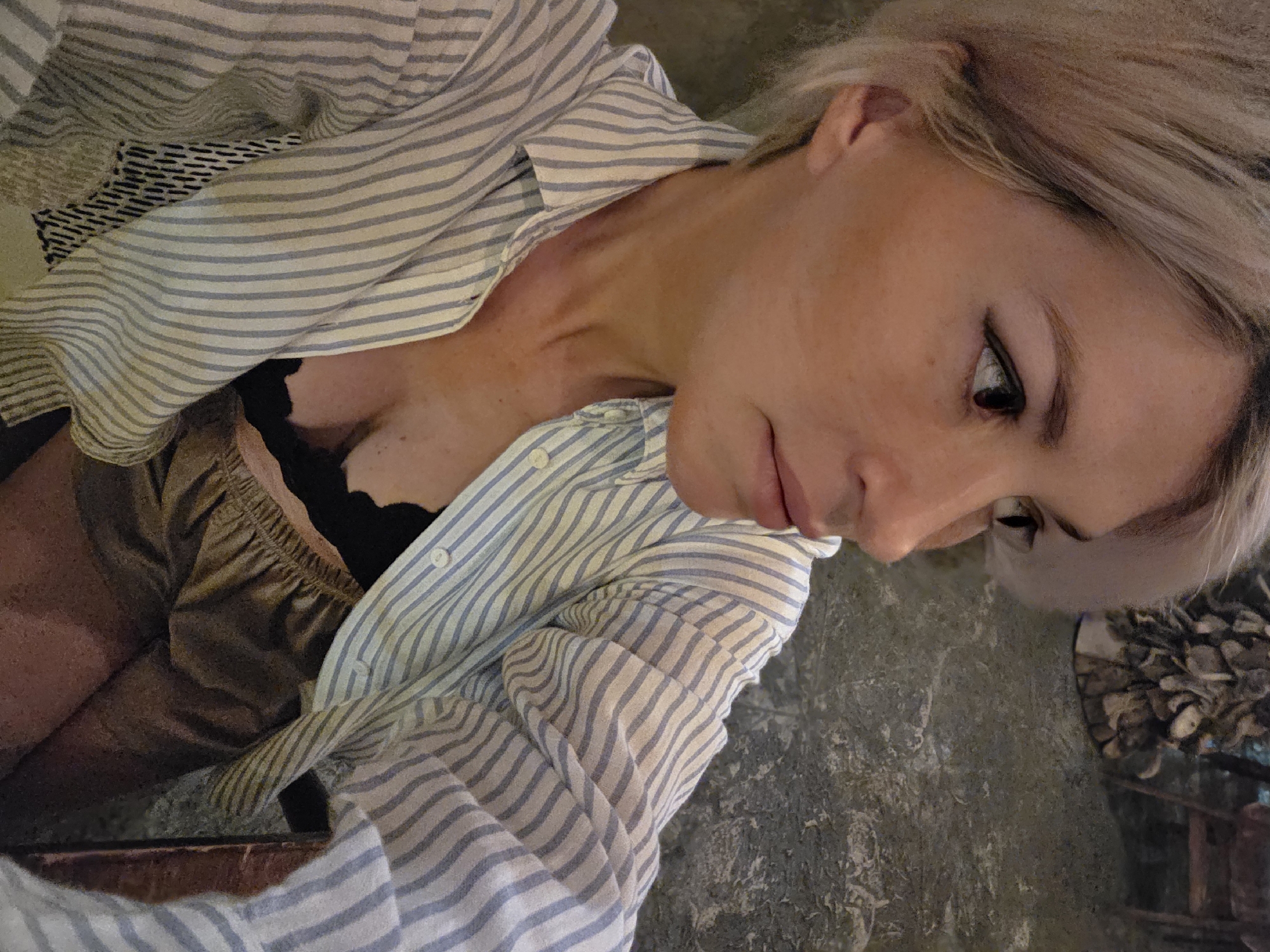 An over-the-shoulder medium shot captures a blonde woman with her head turned to the right, looking away from the camera. She is wearing a light blue and white pinstripe button-up shirt, unbuttoned to reveal a black lace bra and a shimmering gold skirt. Her makeup includes winged eyeliner and subtle lipstick. The background appears to be an indoor setting with textured, grey walls and a hint of foliage or decorative wooden elements to the right. The scene is lit with warm, ambient light. This photograph was taken in North Kuta, Indonesia.