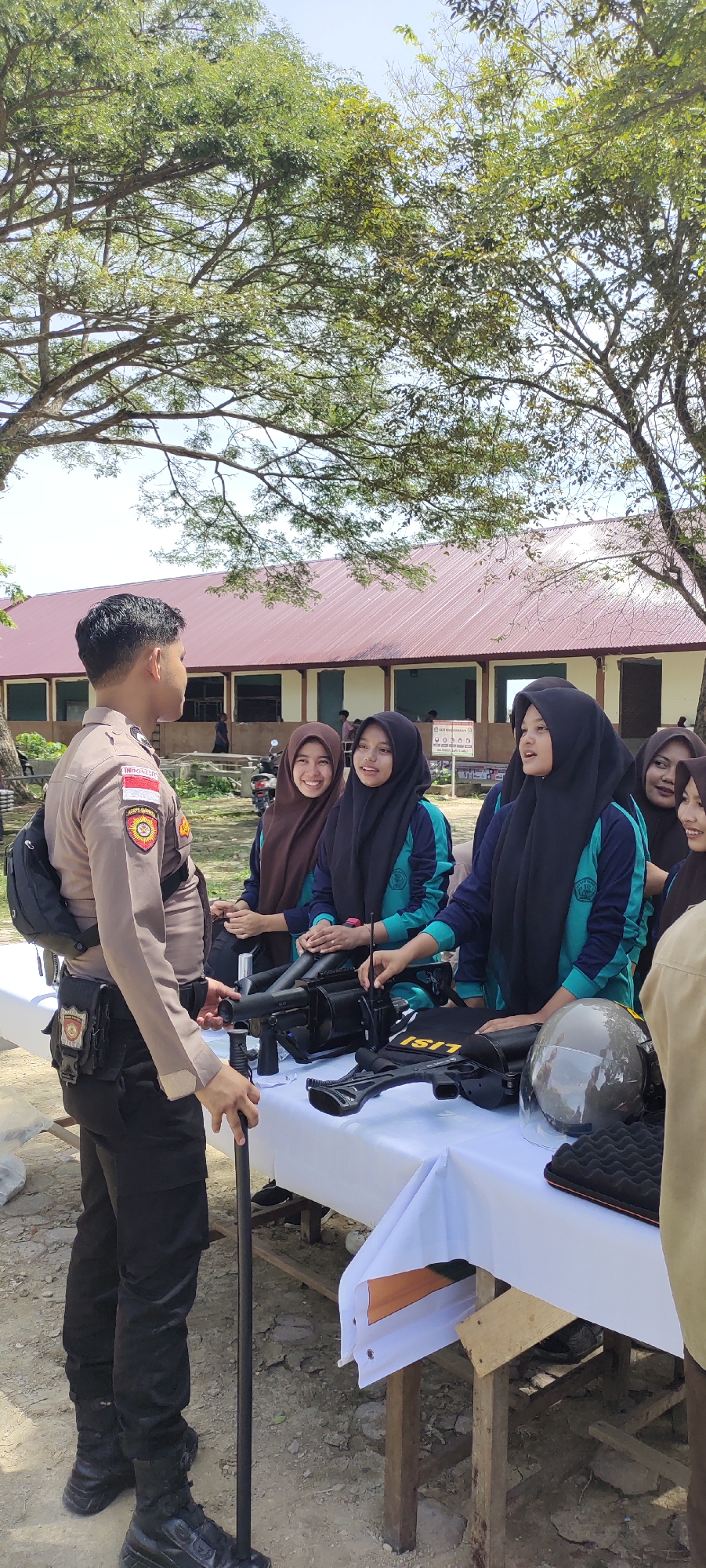 The image captures an outdoor scene in Langnibong, Indonesia, on a sunny day. A male police officer, dressed in a tan uniform with black accents, is standing beside a white-clothed table and addressing a group of five young women, all wearing hijabs and matching teal and dark blue school uniforms. The table is displaying police equipment, including what appears to be a riot shield, helmet, and radio. The women are looking at the items on the table, seemingly engaging with the officer. The background features a one-story building with a red metal roof, and trees overhead.