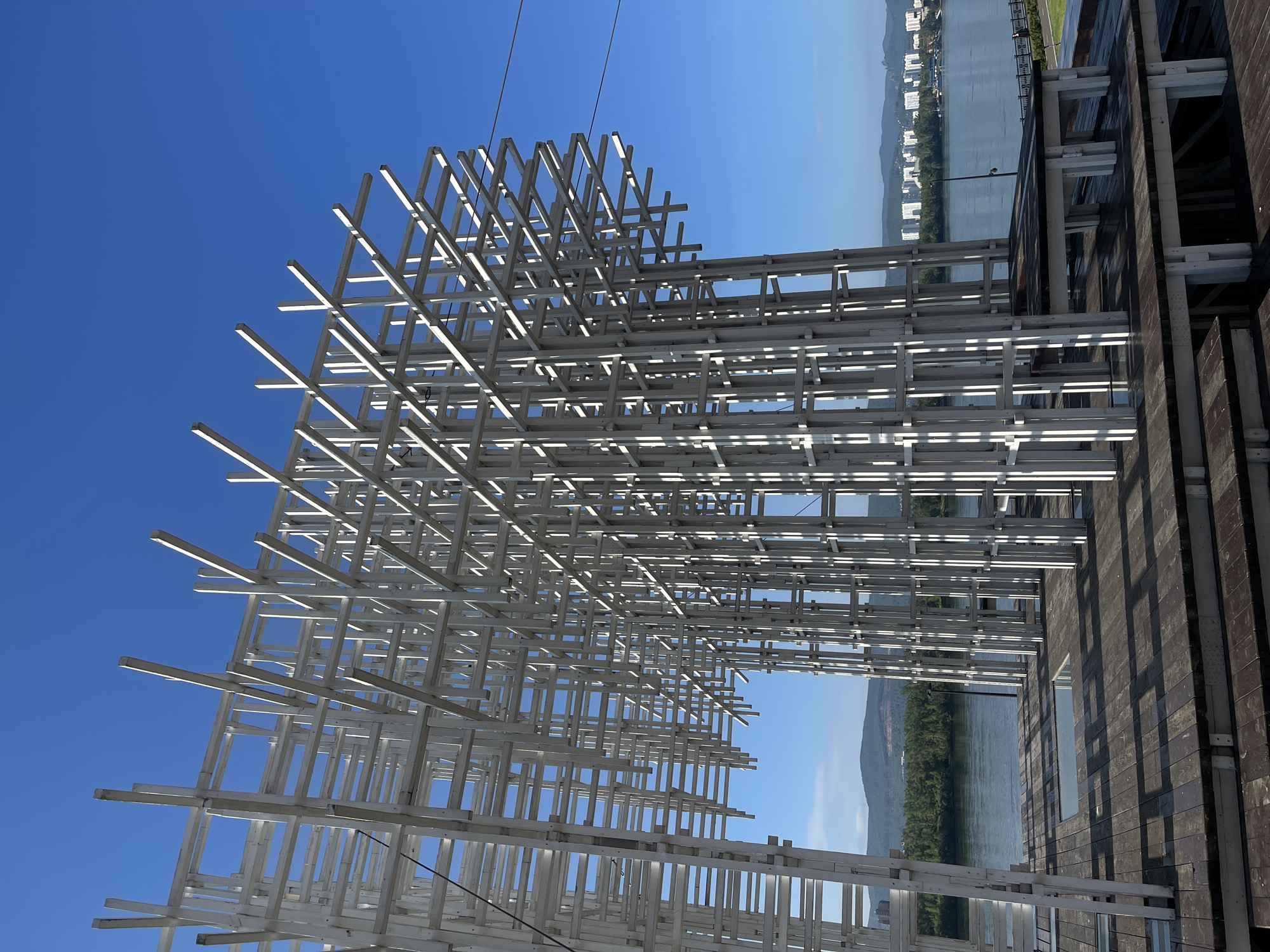 An abstract white geometric sculpture stands on a wooden platform near a wide river. The sculpture is composed of numerous white vertical and horizontal beams, creating a lattice-like structure that extends upwards and outwards. The sky is a clear, bright blue. In the background, across the river, a city skyline with white buildings is visible, nestled against a backdrop of green, tree-covered hills. The wooden platform has a patterned surface created by the shadows of the sculpture's beams. This scene is located in Krasnoyarsk, Russia, at the strelka, where the Yenisei and Angara rivers meet, likely near the Strelka embankment. There are no people or animals visible in the image.