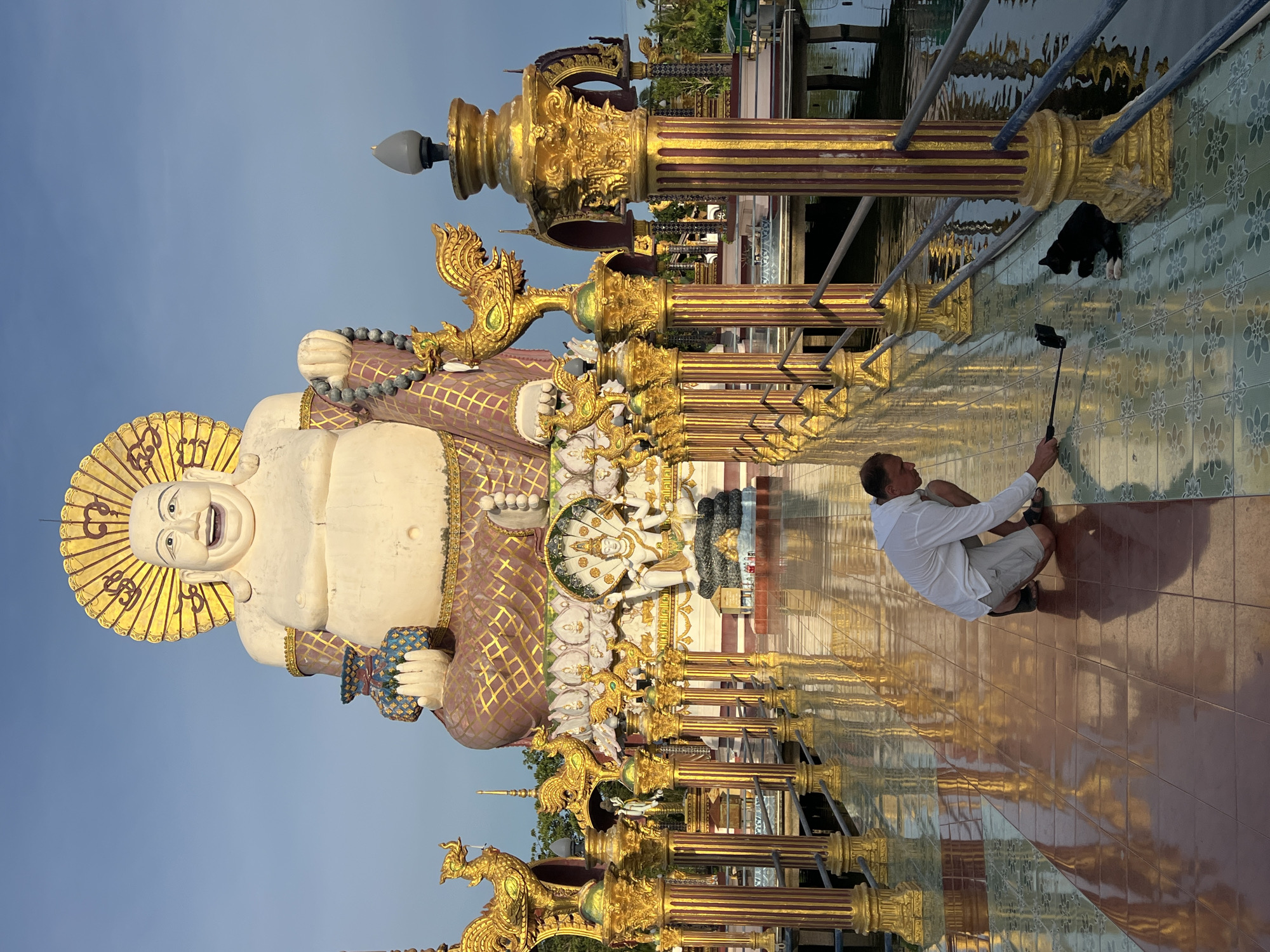 The image depicts a tourist taking a selfie with a large, white statue of a laughing Buddha in Baan Plai Laem, Thailand. The statue is adorned with ornate gold embellishments and a halo. The tourist, a man wearing a white shirt and shorts, is crouching down to capture the photo with a selfie stick. In the background, a black cat is visible perched on a golden railing. The scene appears to be set outdoors, with a clear blue sky suggesting it is daytime. The ground is tiled with a reflective surface, mirroring the golden statues and creating a vibrant, sun-drenched atmosphere. The overall mood is one of joy and exploration, typical of a tourist experiencing a cultural landmark. The architecture and the statue itself strongly suggest a religious or cultural site, likely a temple or shrine, given the context of Thailand. There is no visible text in the image.