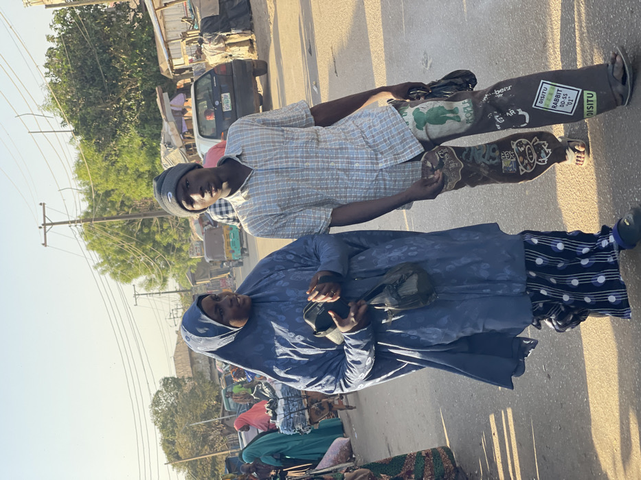 Two individuals are the focal point of this outdoor snapshot. The person in the foreground wears a blue hijab-like garment with a pattern and carries a black bag. Behind them, a younger person sports a checkered shirt, a Nike beanie, and unique lower garments covered in drawings and stickers. They are positioned on a road, likely a bustling area, as evidenced by the surrounding vehicles and pedestrians. The backdrop features buildings, utility poles, and vegetation, suggesting an urban or semi-urban setting.