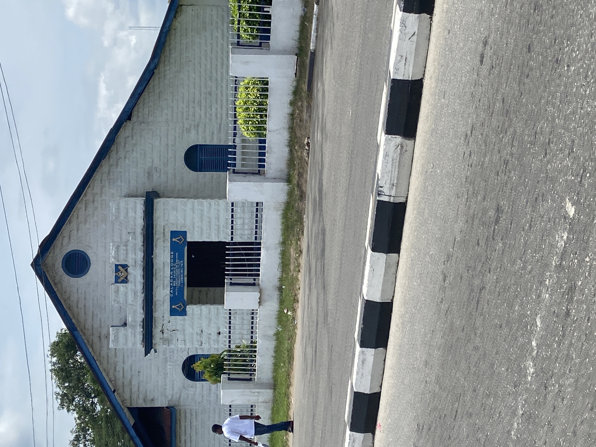 The image shows the Calabar Lodge No. 3416 E.C. Masonic lodge, a white building with blue trim, in Calabar, Nigeria. A man in a white shirt and jeans walks past the building. A black and white curb separates the building from the gray asphalt street. The sky is visible in the background.